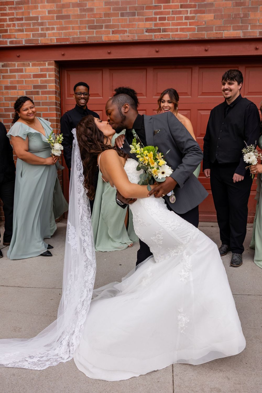 Bride and groom share a romantic dip kiss surrounded by wedding party in sage green dresses outside brick building.