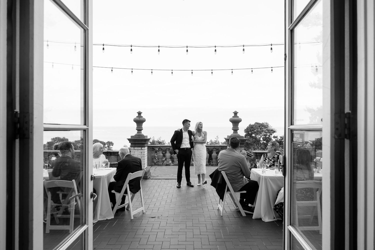 Black and white photo of a couple standing on an outdoor terrace during a wedding reception with string lights above.