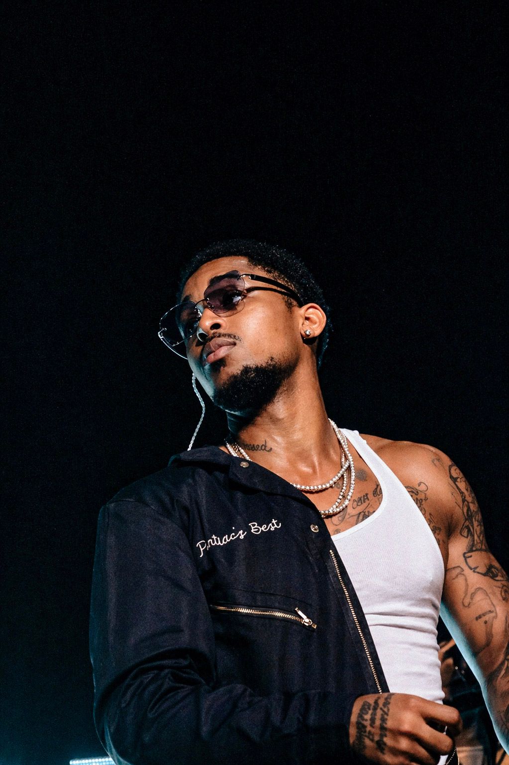 Young man with tattoos, sunglasses, and chain necklace wearing a white tank top and dark jacket performing on stage.