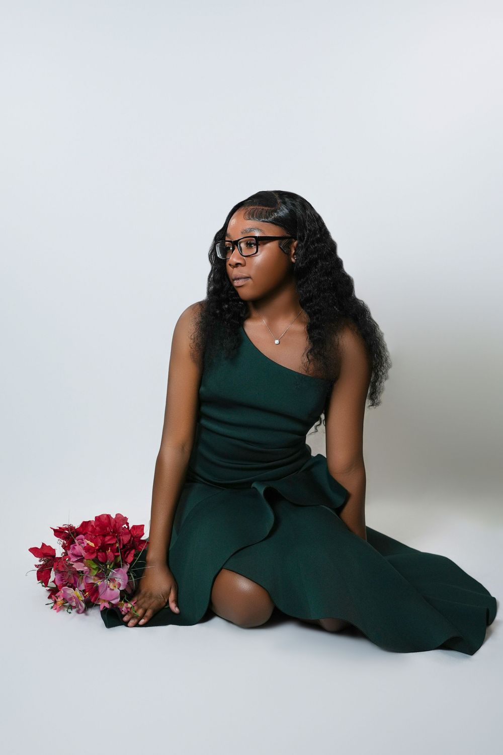 A young Black woman in a green one-shoulder dress sits on white floor holding pink flowers, wearing glasses.