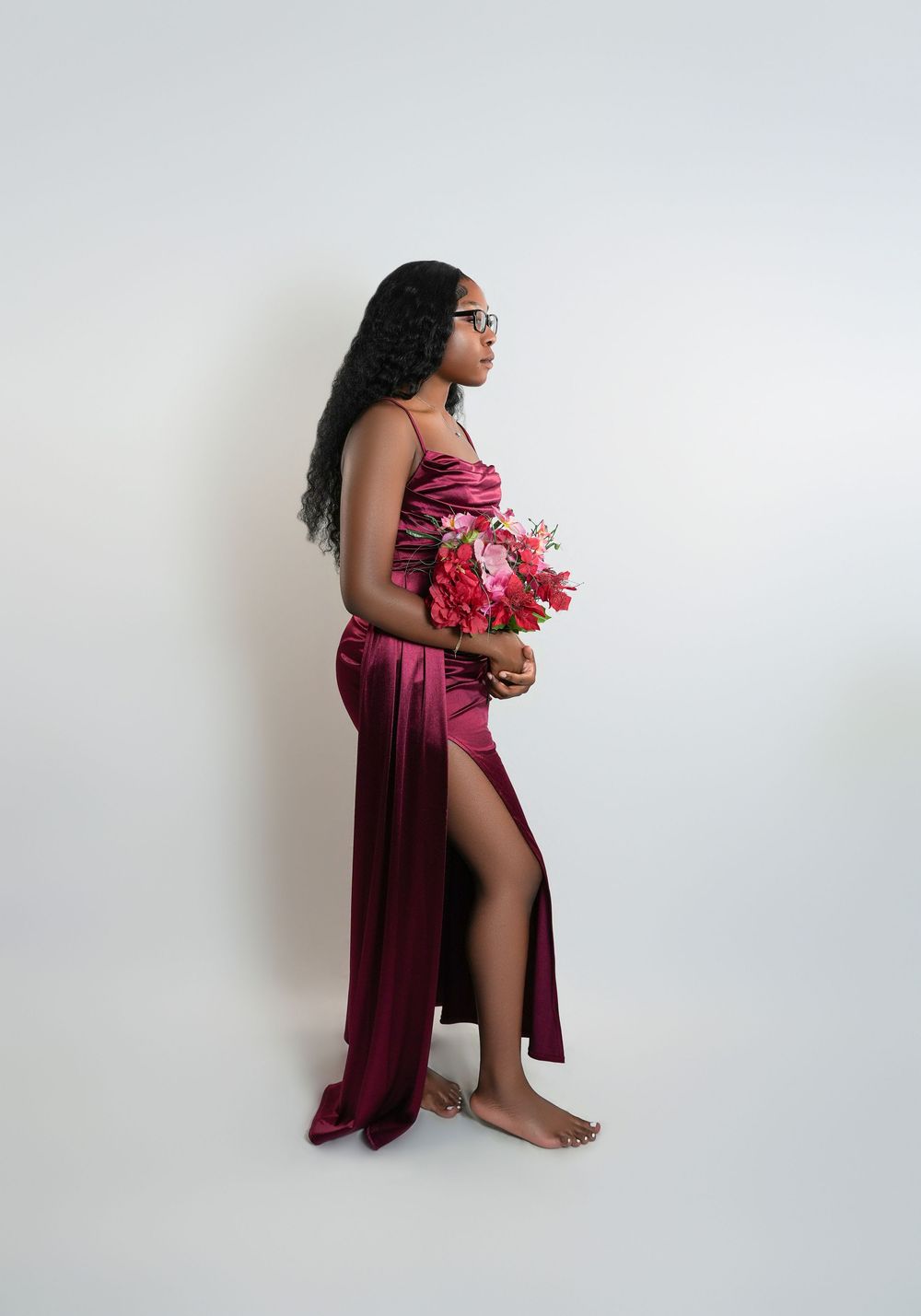 Young woman in burgundy satin gown with slit holds pink floral bouquet, posing sideways in studio.