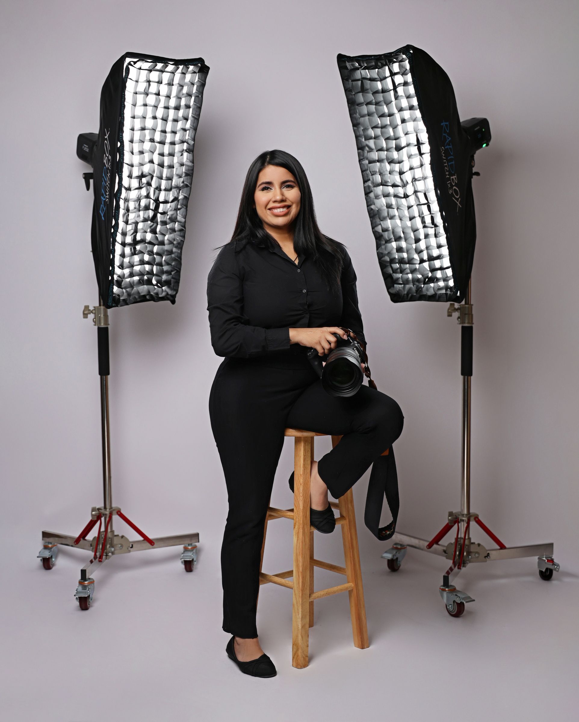 Female photographer in black outfit sits on stool between two studio softbox lights holding a camera.