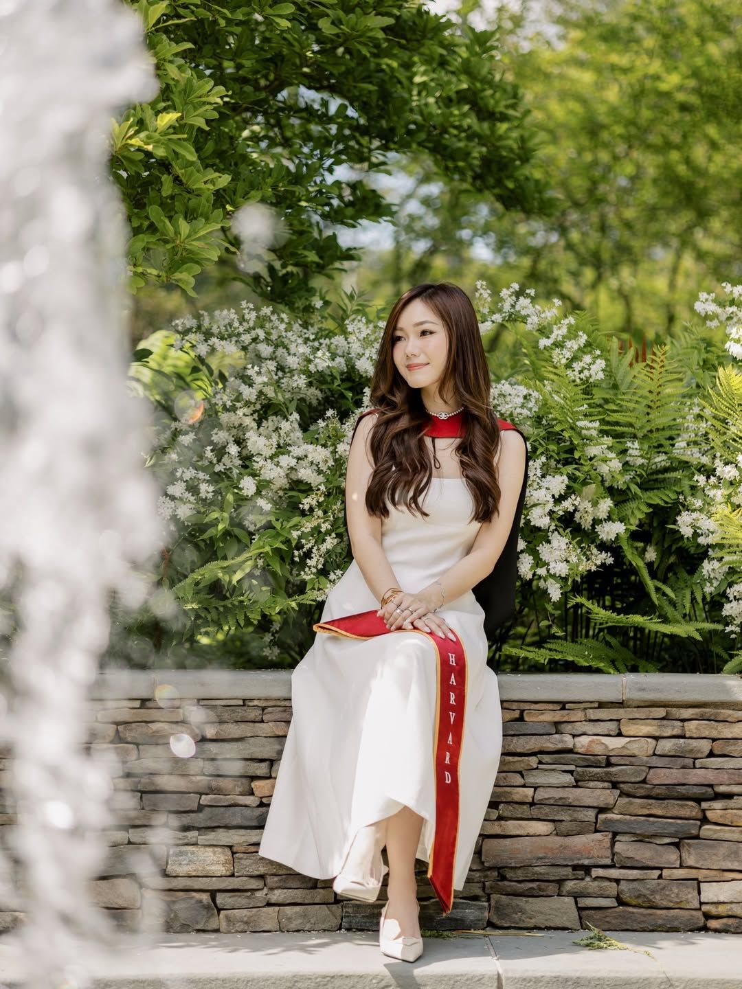A graduate in black gown and red sash sits on a white bench holding flowers, with autumn leaves falling near a modern building.