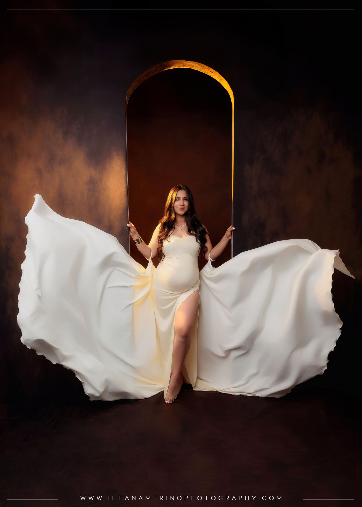 Pregnant woman in flowing white dress with wings spread in golden arch studio setting.