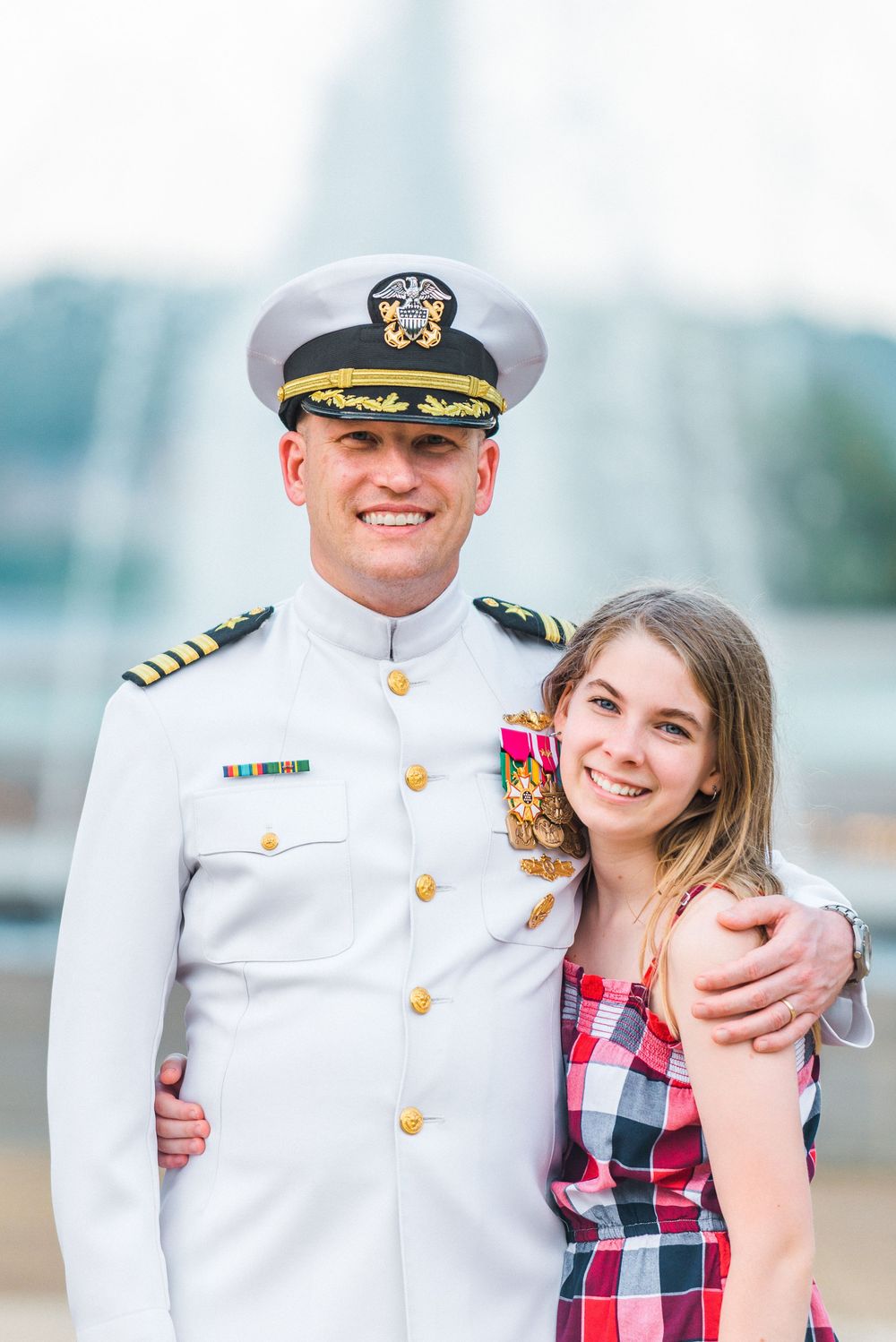 A naval officer in dress whites stands with companion in a plaid dress.
