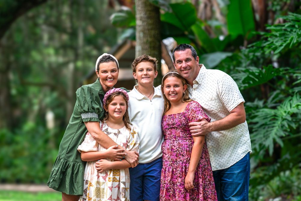 A happy family of five poses together outdoors in a lush tropical garden setting for a professional portrait.