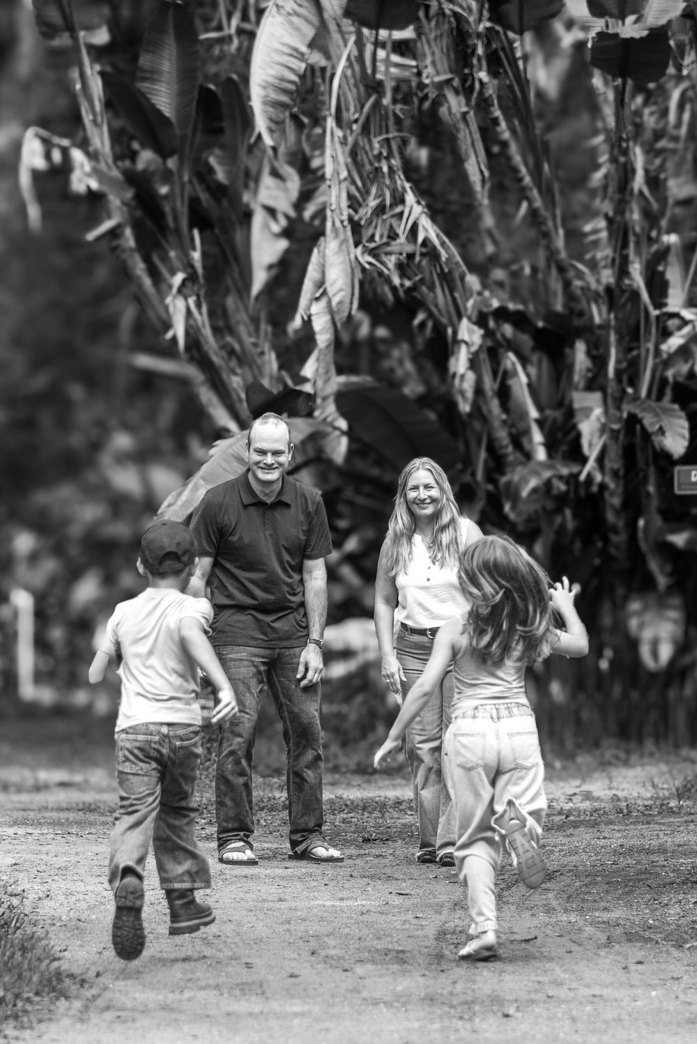 Black and white photo of a family with two children running toward smiling parents on a tree-lined path.