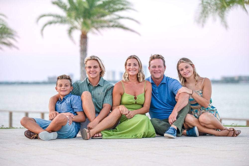 Family of five smiling together on a beachside boardwalk with palm trees and ocean in the background.