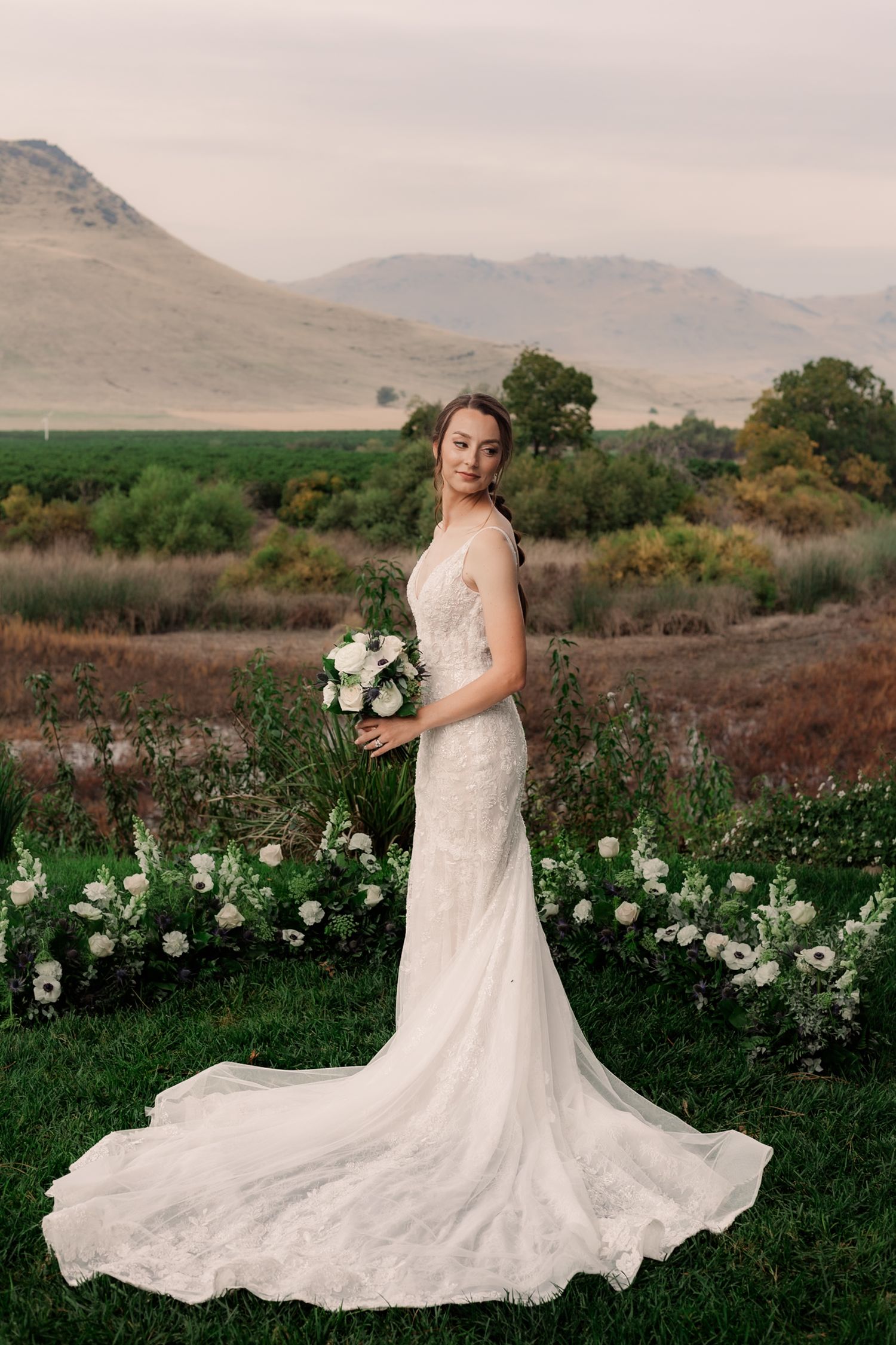 A white wedding dress with a flowing train is displayed against a backdrop of mountains and white flowers in a field.