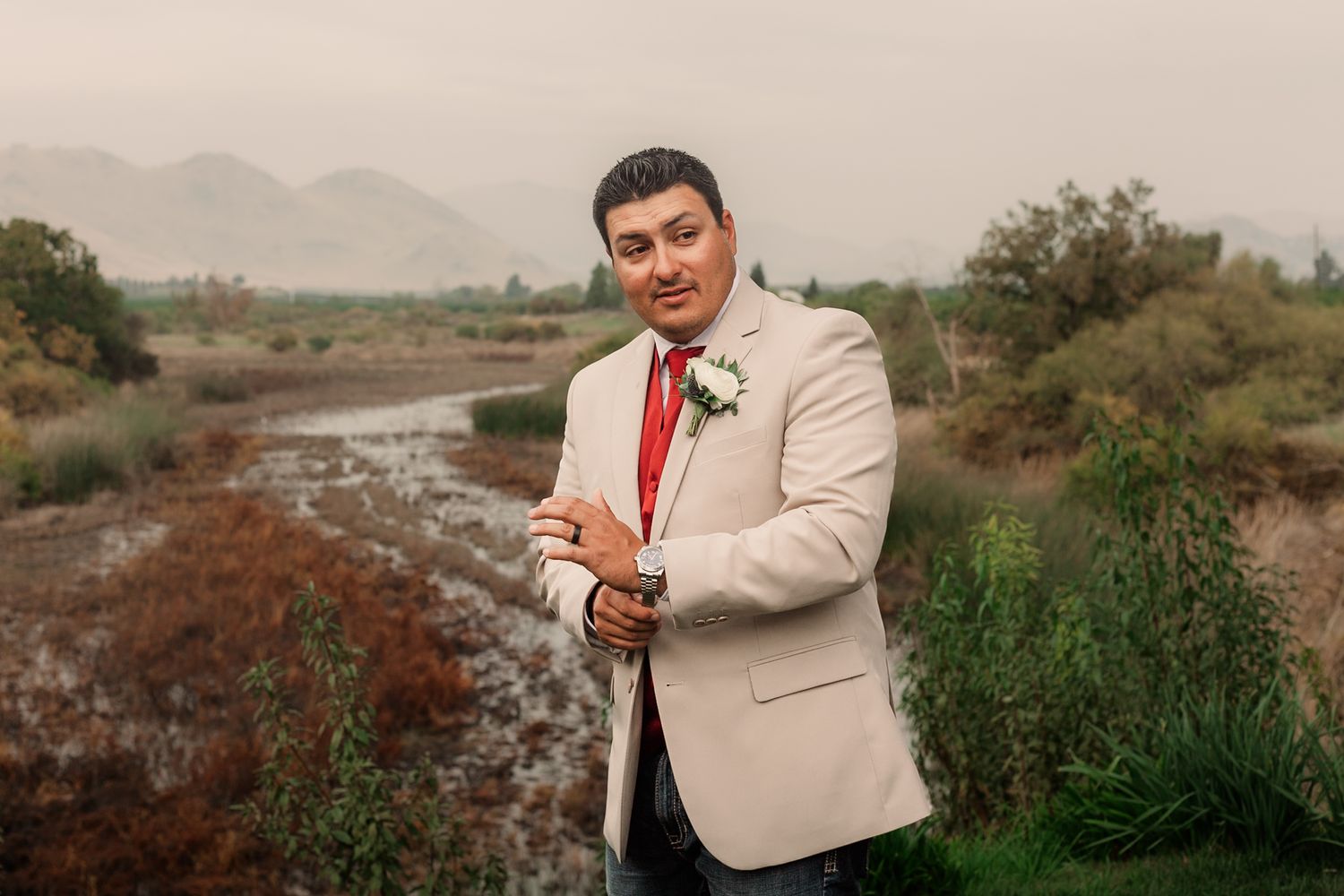 A person in a beige blazer and red tie poses in front of a desert landscape with mountains and brush.