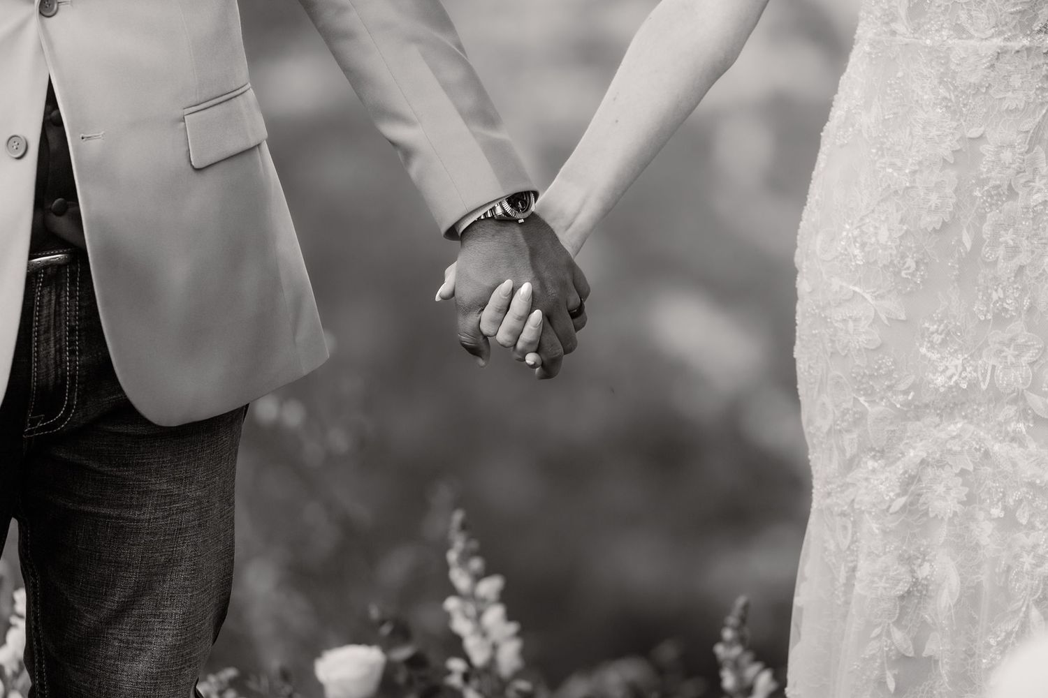 A couple holds hands during an outdoor wedding ceremony while wearing formal attire and standing in a garden setting.