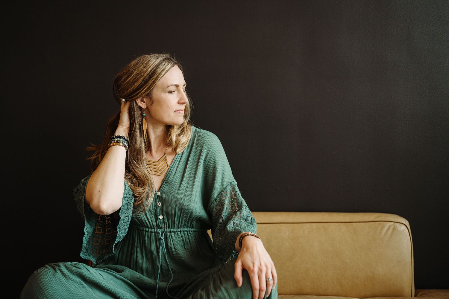 Person in green shirt sits thoughtfully on tan couch while looking away from camera in moody lighting.