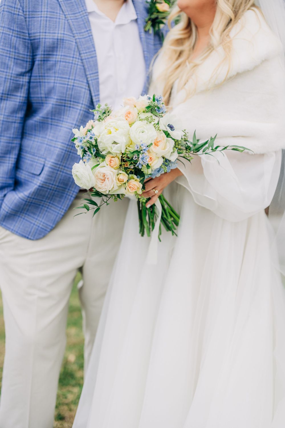 A wedding couple poses outdoors, showing details of a blue checkered suit jacket and white dress with a peach floral bouquet.