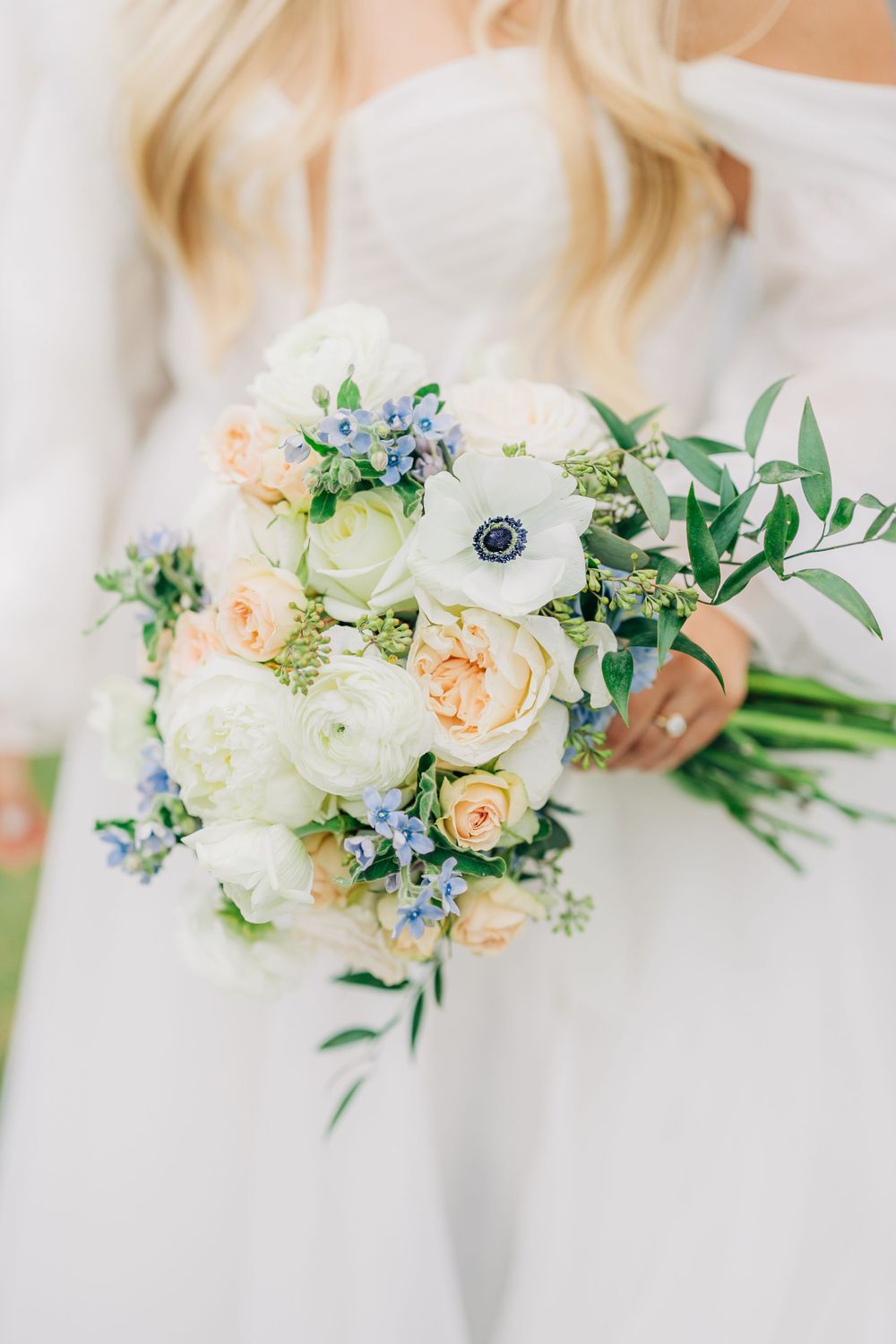 Delicate bridal bouquet with peach garden roses, white flowers, and blue forget-me-nots arranged with green foliage.