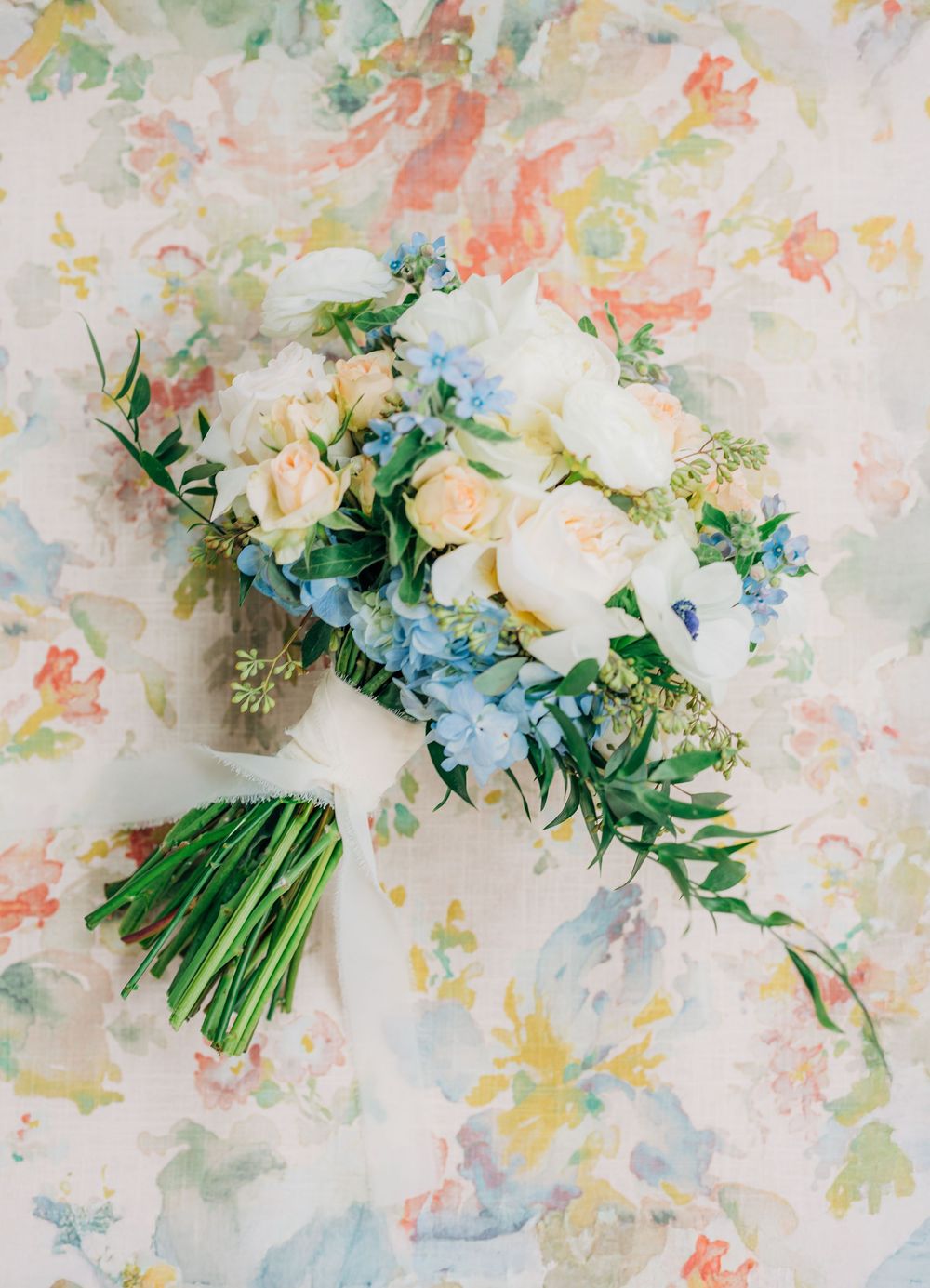 A delicate bridal bouquet featuring white roses, blush peonies, and blue delphinium flowers against a pastel watercolor backdrop.