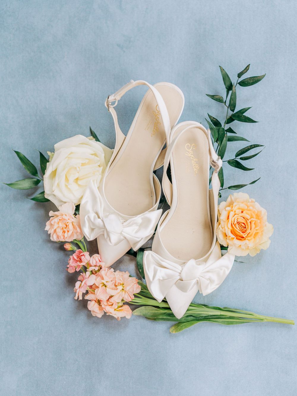 Elegant white bridal shoes paired with peach roses and white floral arrangements on a soft blue background.