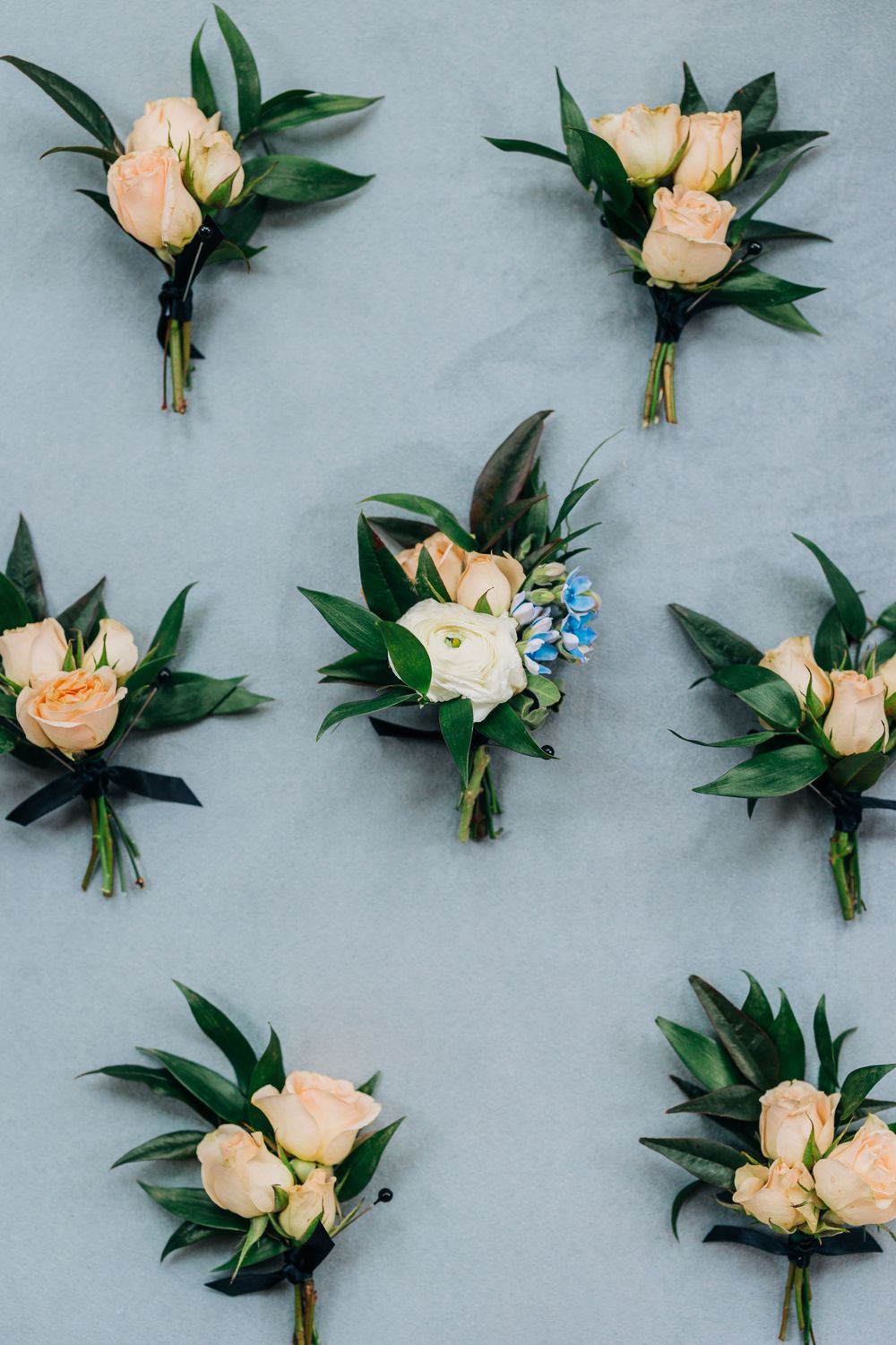 Peach-colored rose boutonnieres with green leaves arranged in a row on a light gray background.
