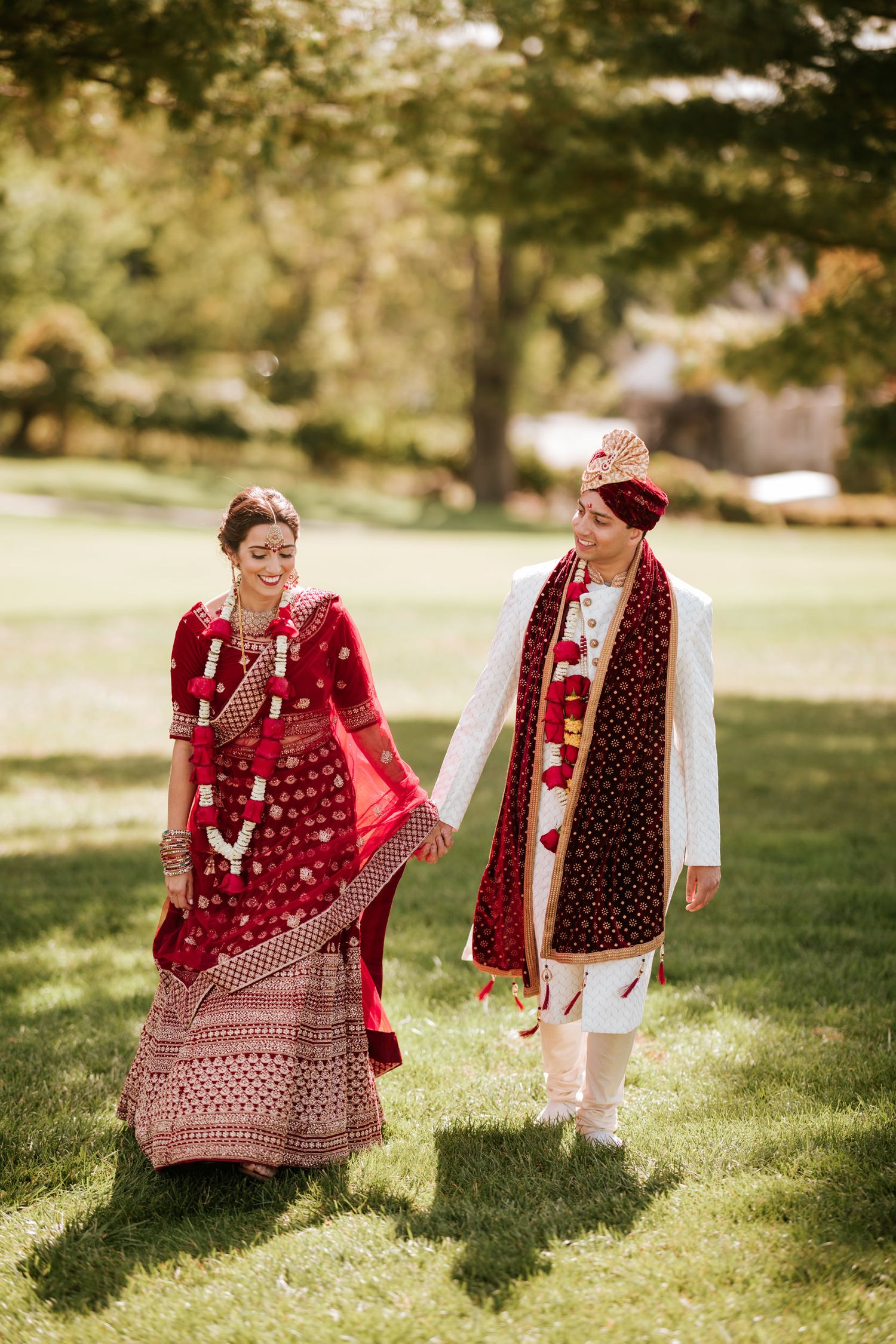 A couple in traditional South Asian wedding attire share playful moments on a sunlit grassy lawn.
