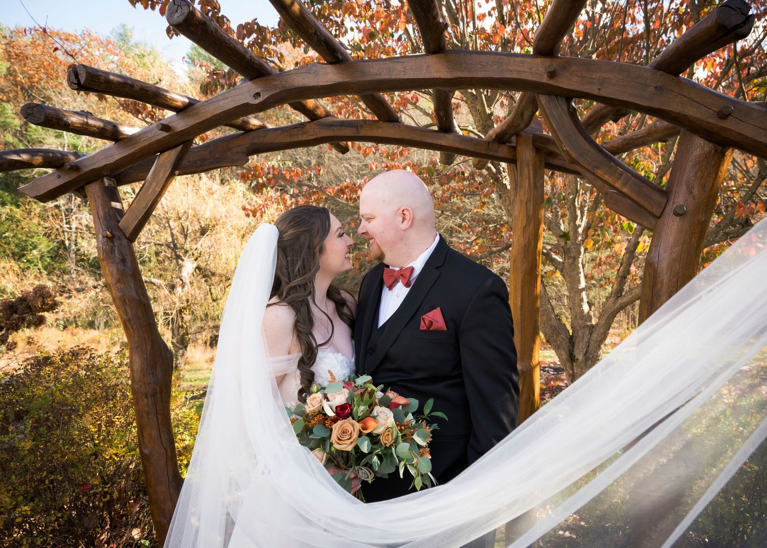 A romantic autumn wedding scene under a rustic wooden arbor with flowing white veil and autumn-colored bouquet.