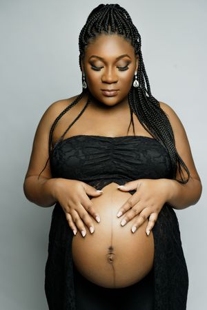Elegant maternity photograph featuring expecting mother in black dress with braided hair against neutral backdrop.