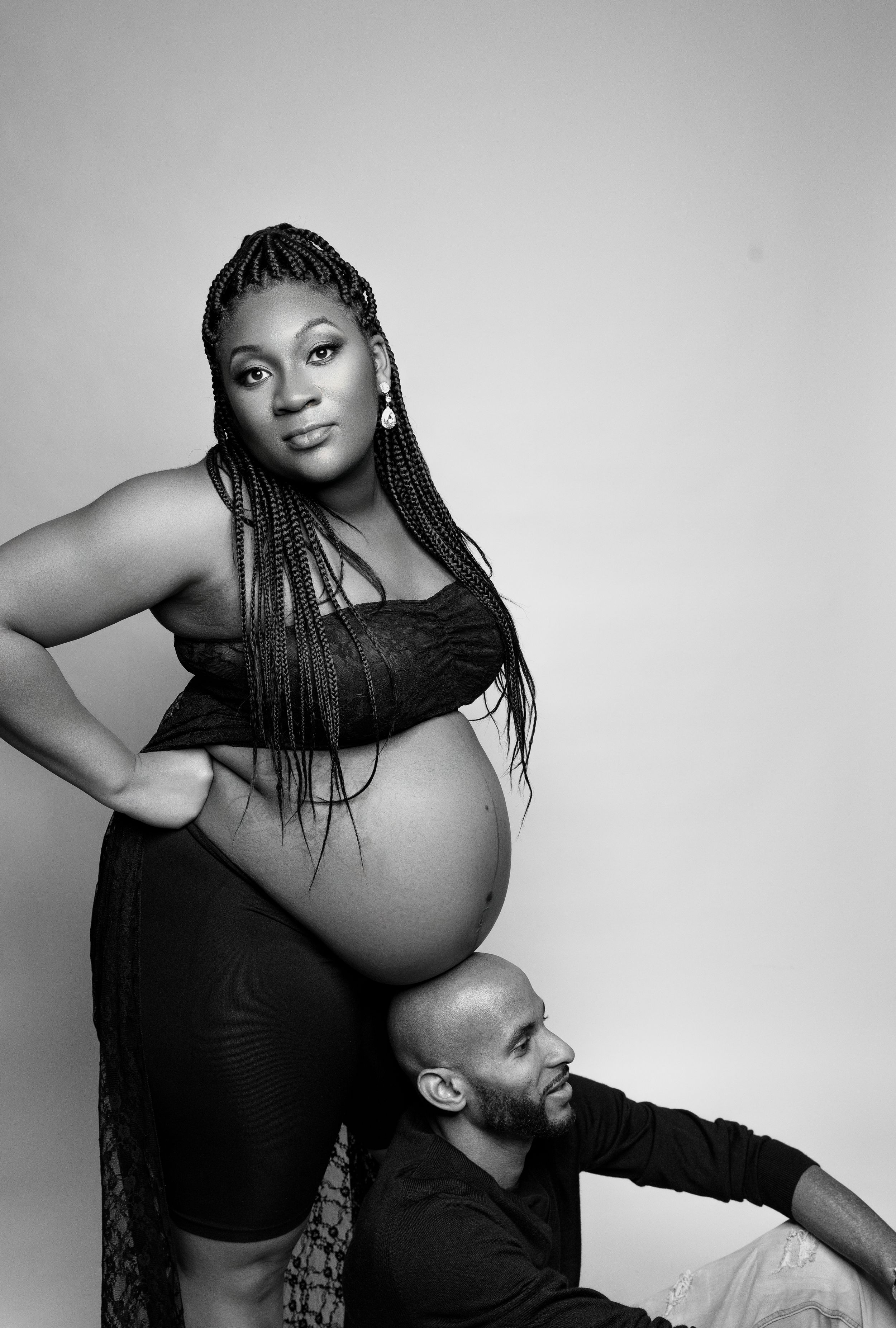 Black and white maternity photo featuring an expectant mother in dark clothing with long braids posing with partner.