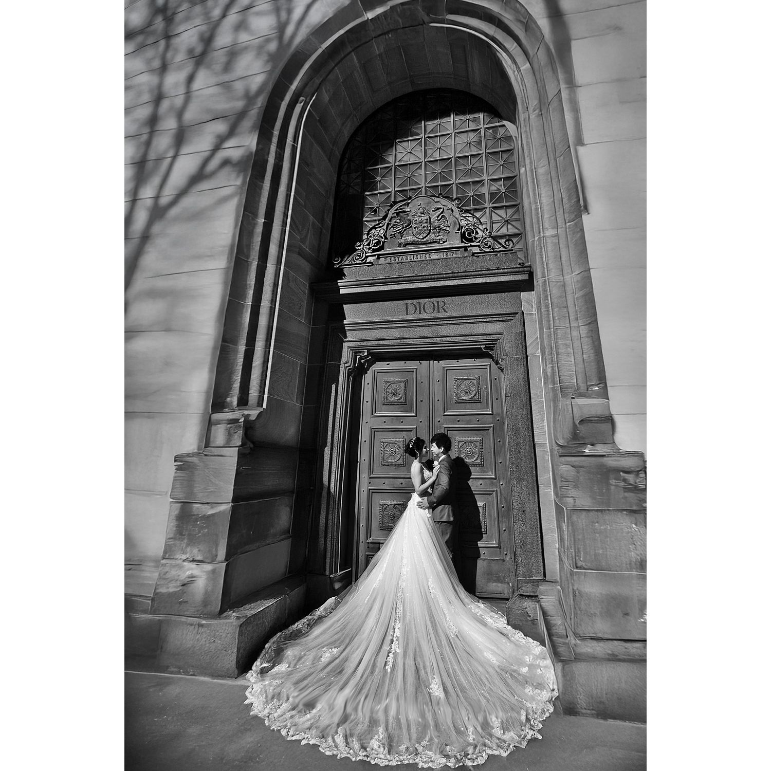 Bride and groom share a romantic kiss before grand arched stone church doors in black and white photo.