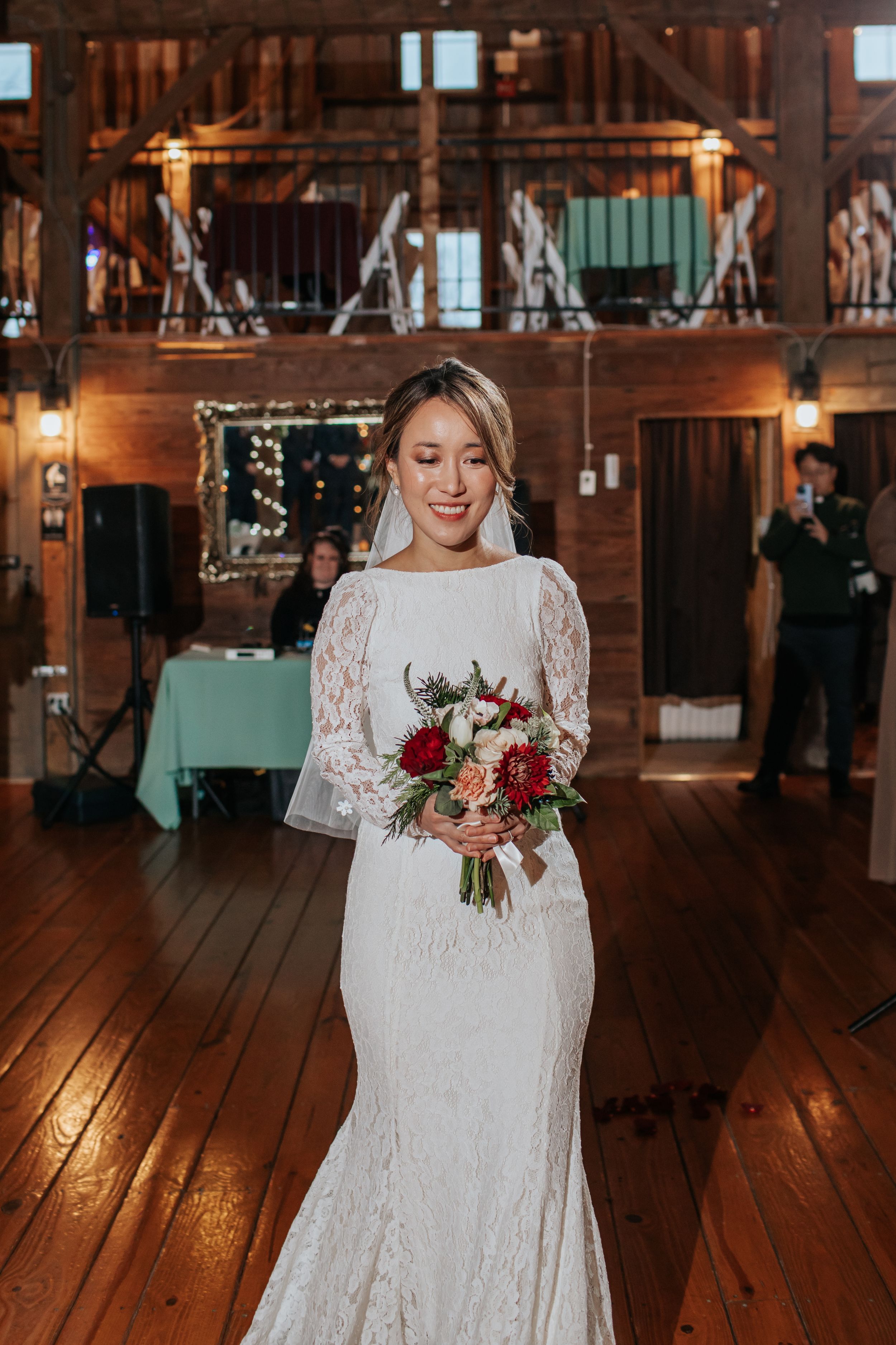 A bride in a lace wedding dress holds a vibrant red and pink bouquet in a rustic wooden venue.