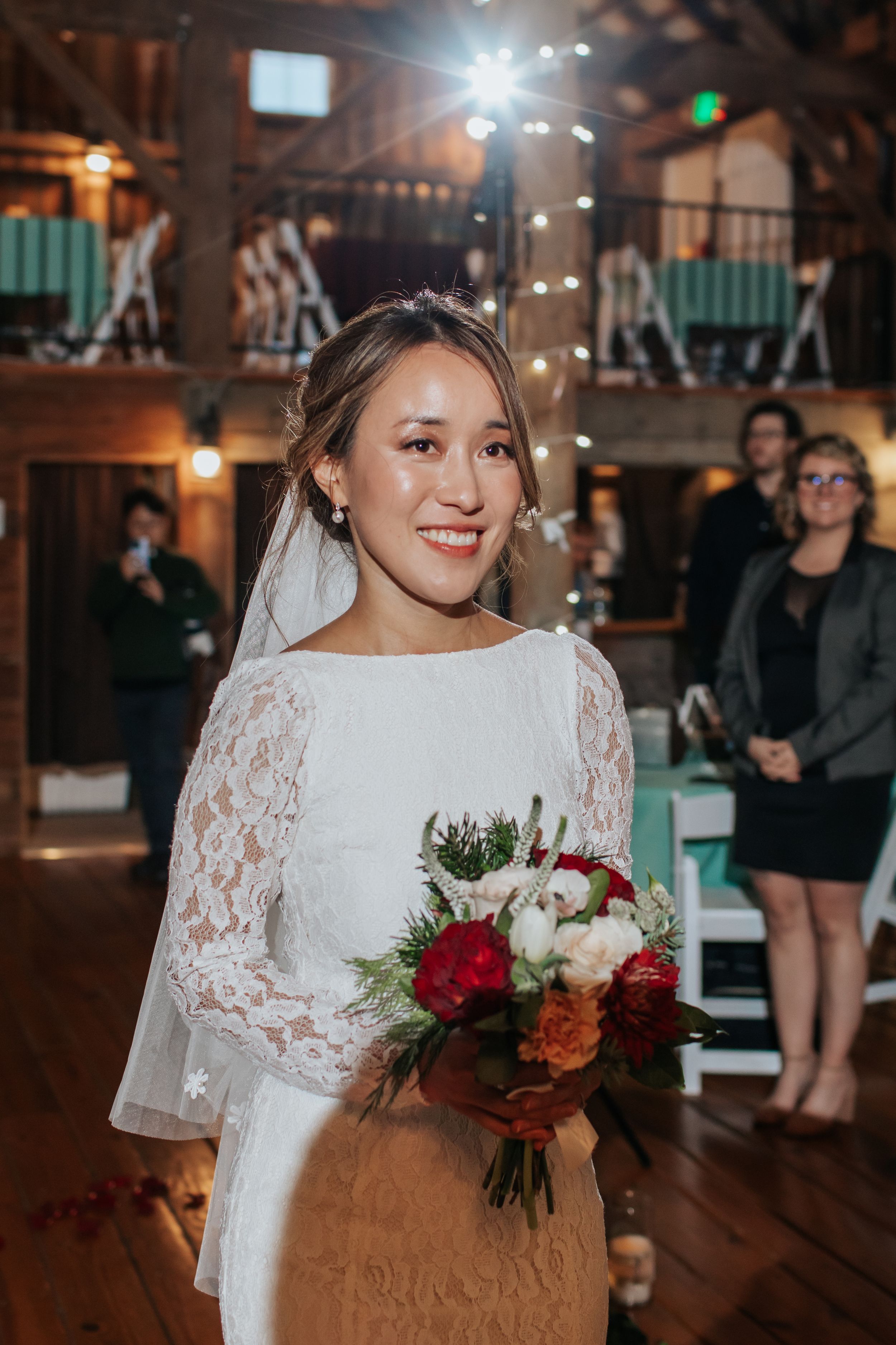 A bride in a white lace wedding dress holds a vibrant red and white bouquet in a rustic wooden venue.