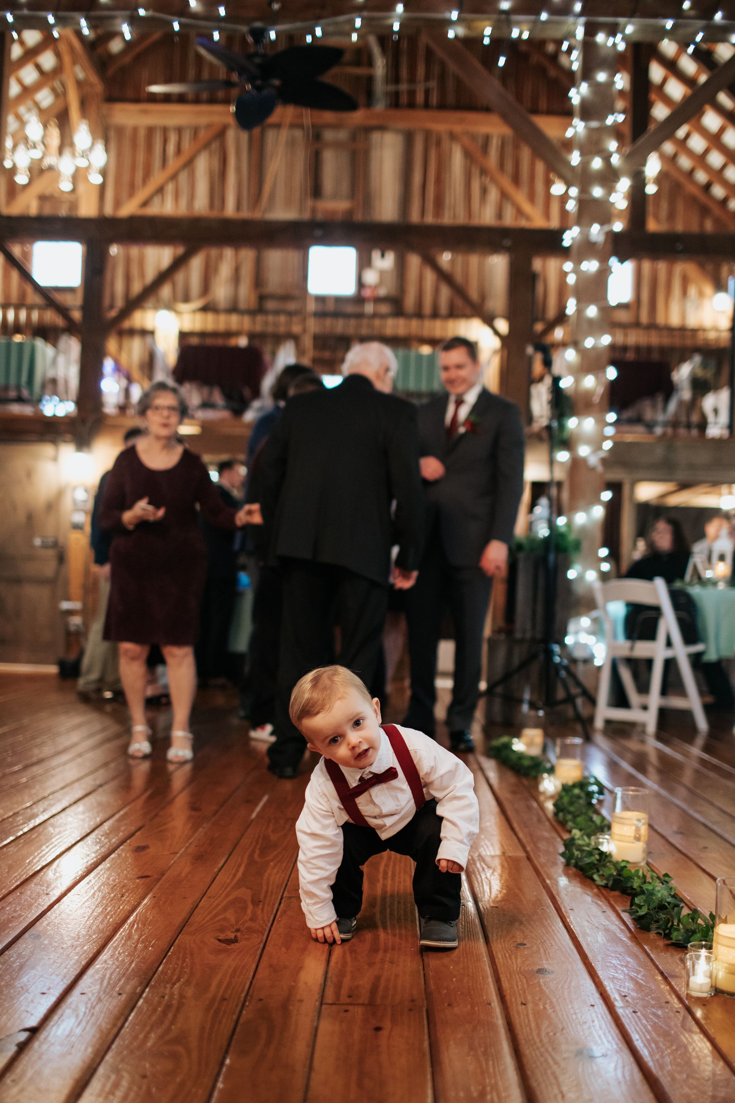 A young ring bearer in formal attire crawls playfully across a wooden barn floor during a wedding.