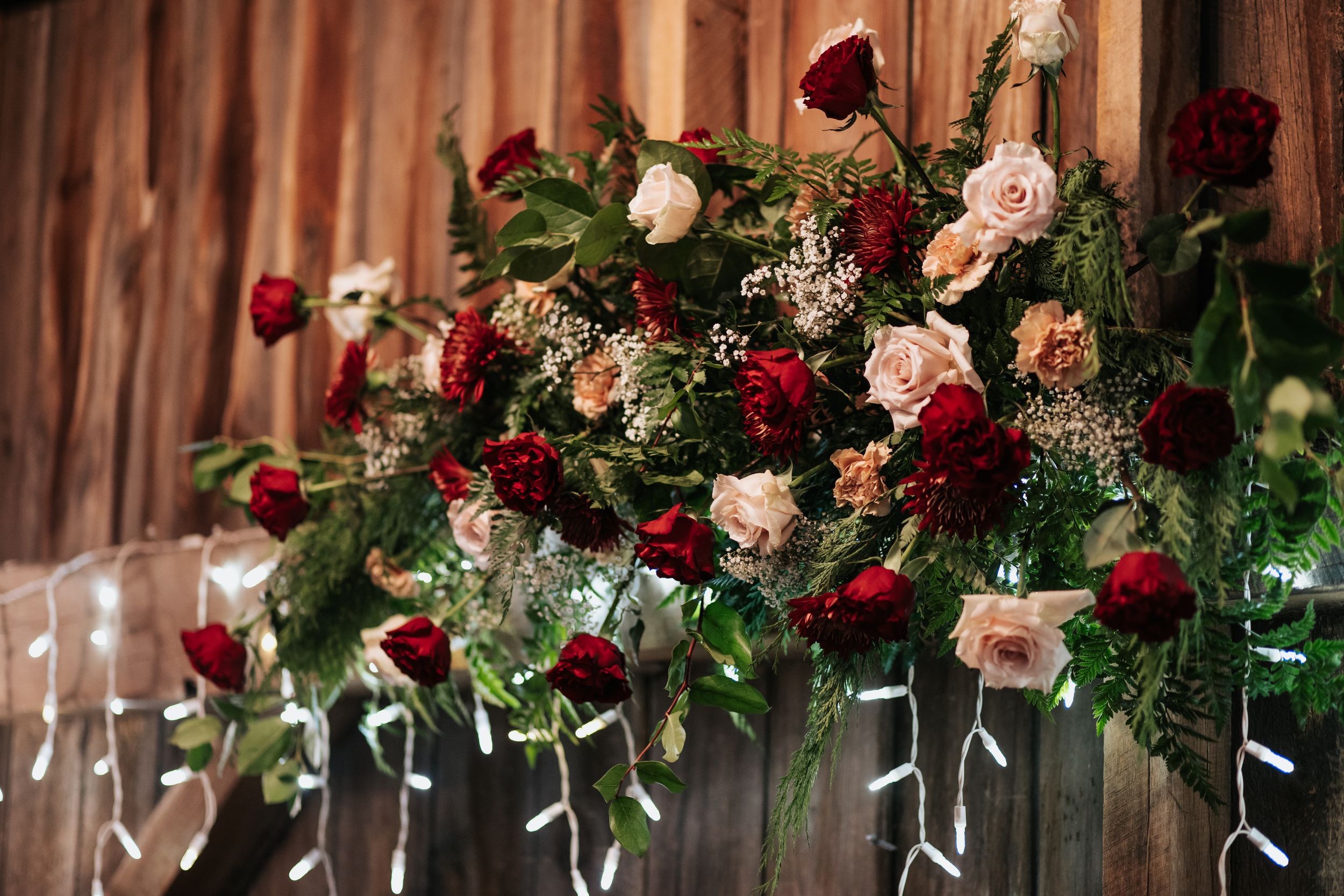 Elegant floral arrangements with red and pink roses hang from wooden beams in a rustic wedding venue.