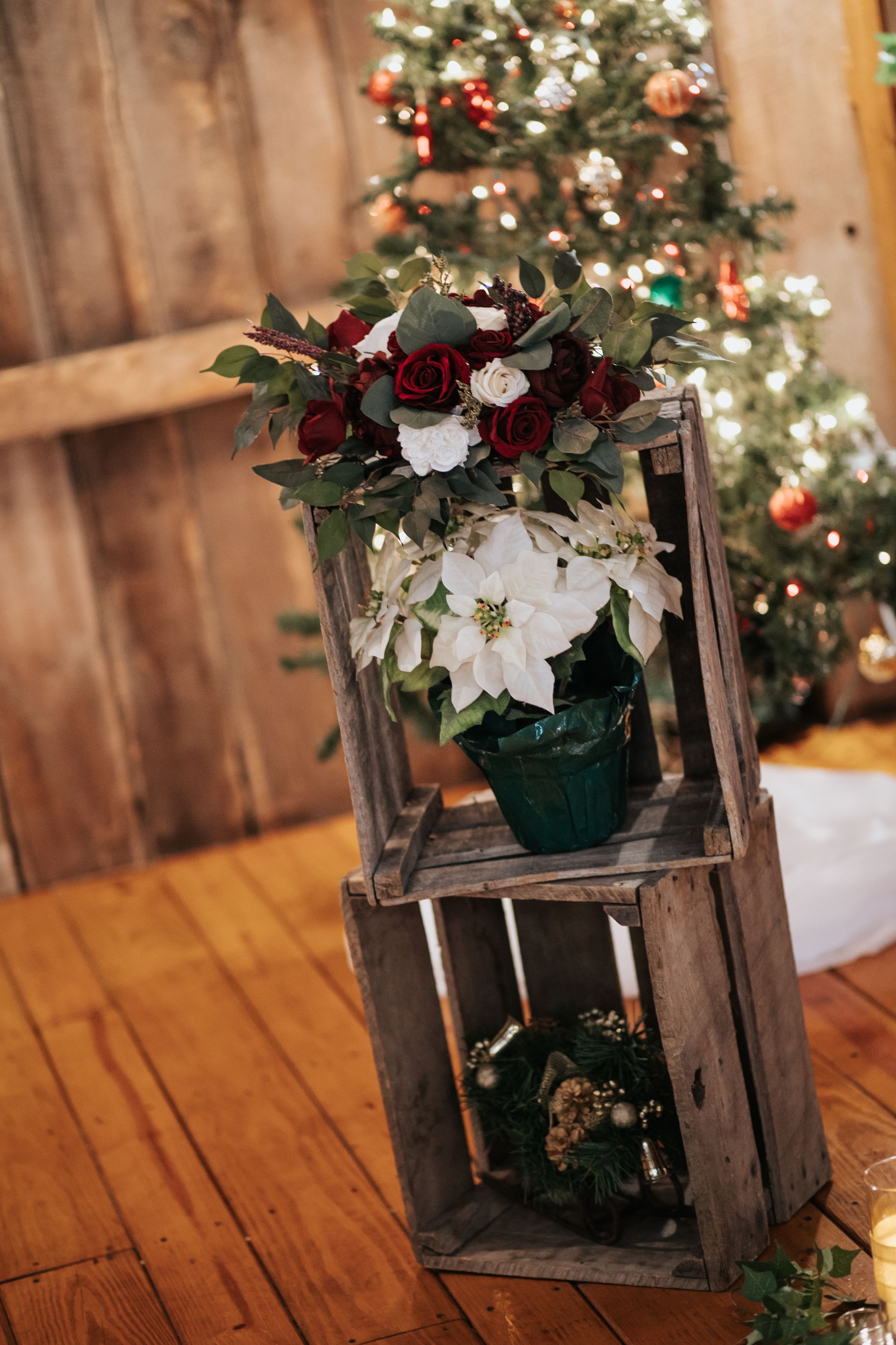 Rustic wooden crate display with white poinsettias and red floral arrangement in front of decorated Christmas tree.
