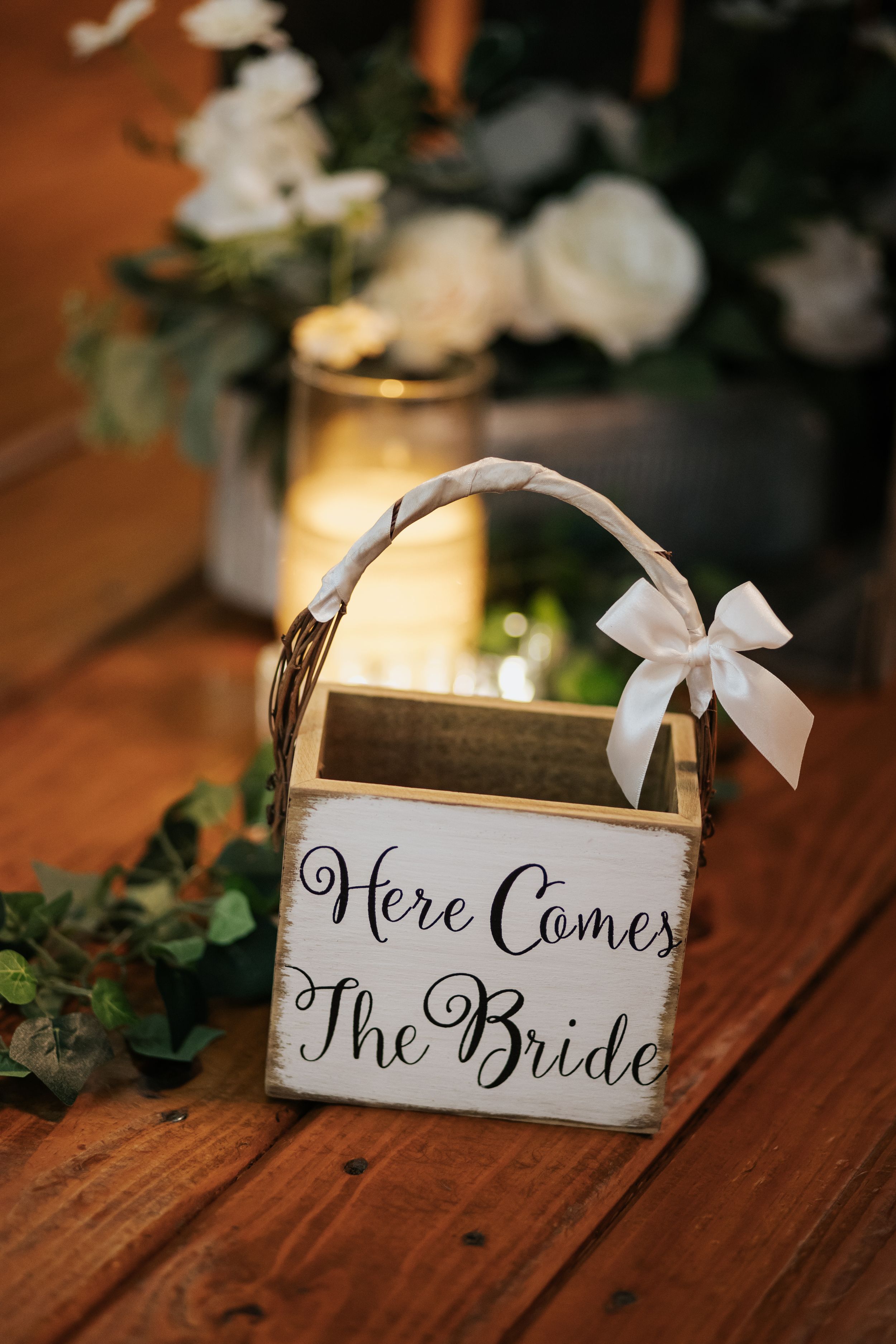 Wedding signage reading 'Here Comes The Bride' displayed on rustic wooden surface with greenery and candlelight.