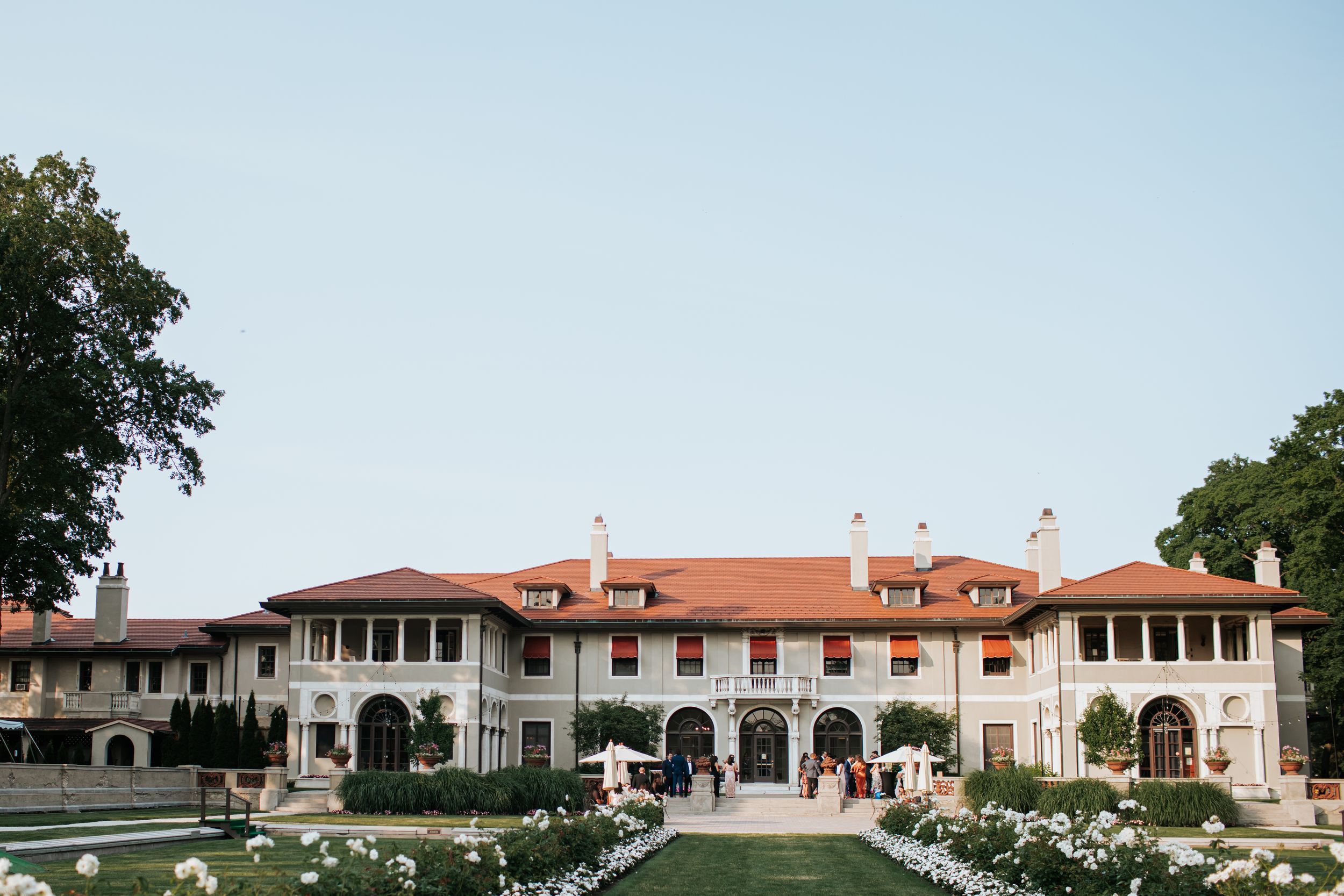 A grand Mediterranean-style mansion with terracotta roof and white archways surrounded by manicured gardens.