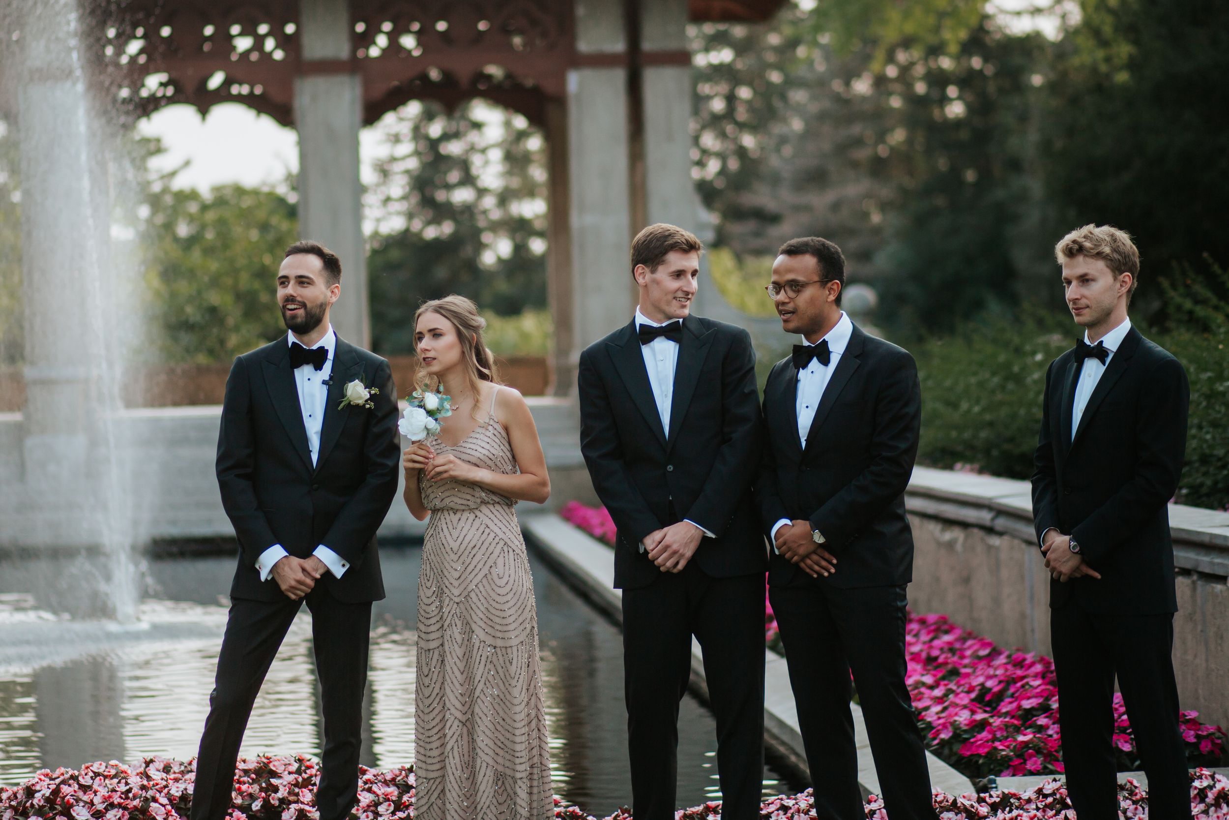 Wedding party members in formal attire stand together by a fountain with pink flowers in the background.