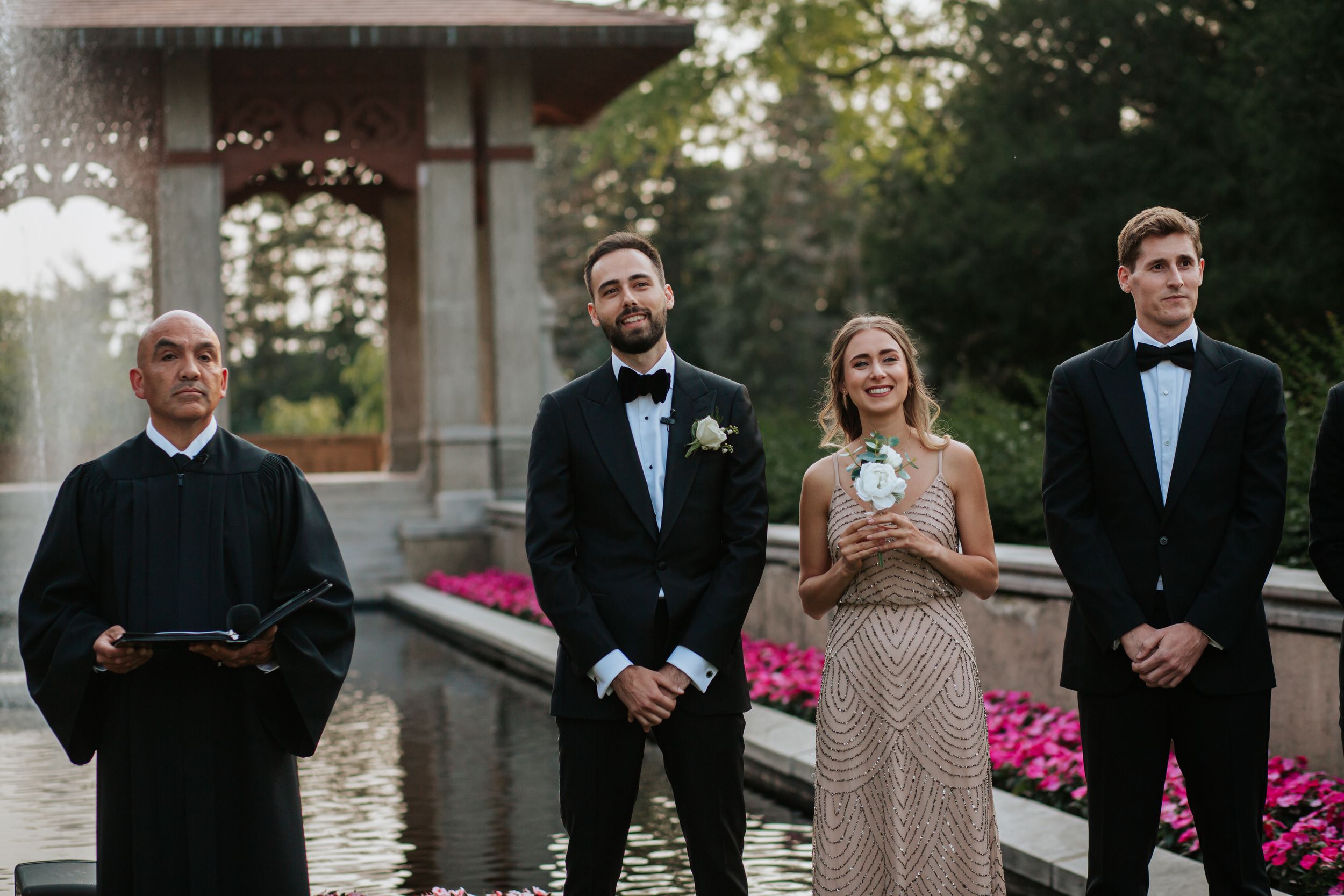 Wedding party members stand in formal attire on a stone bridge near pink flowers and a pond.
