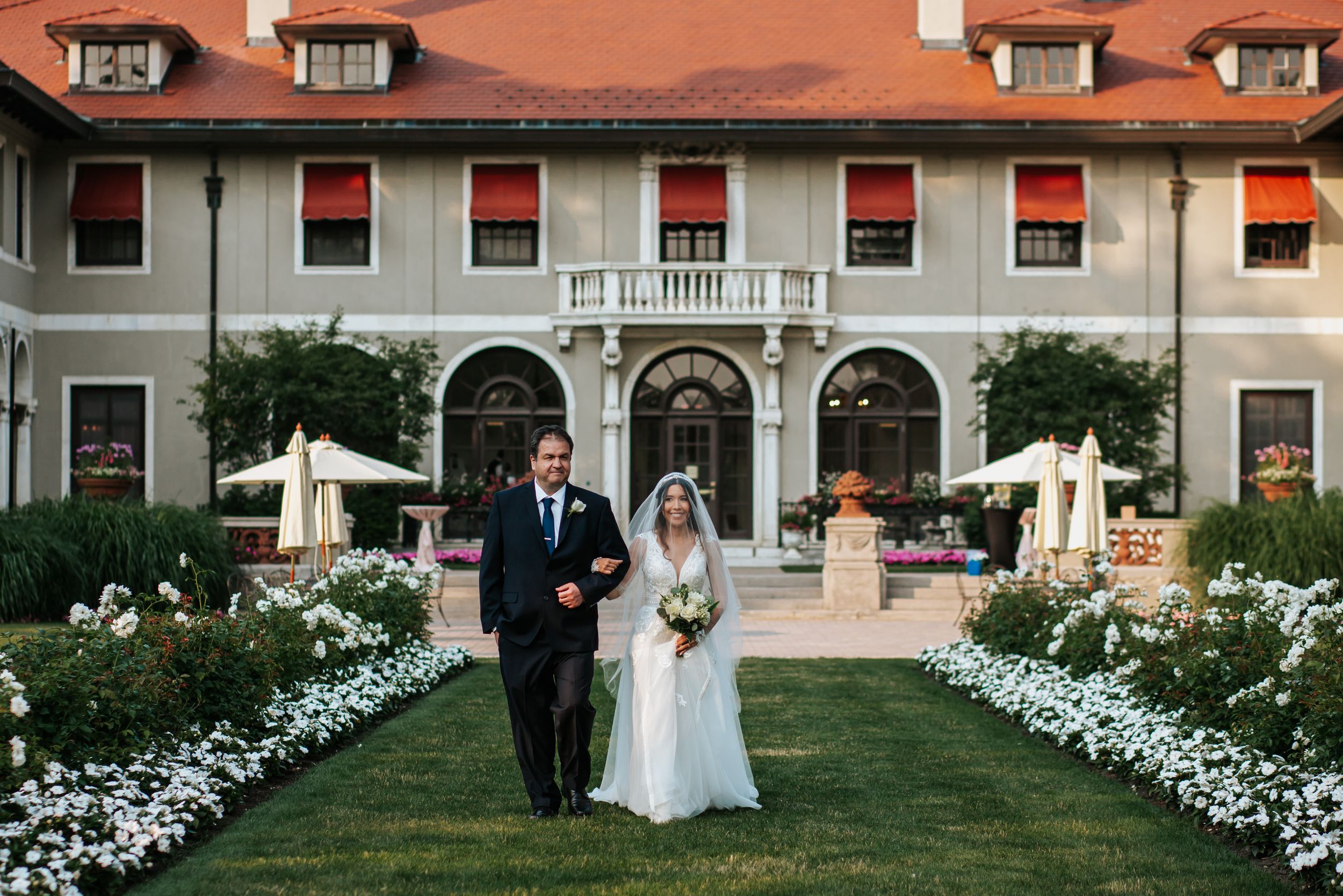 A couple in wedding attire walks down a garden path lined with white flowers in front of an elegant mansion with red awnings.