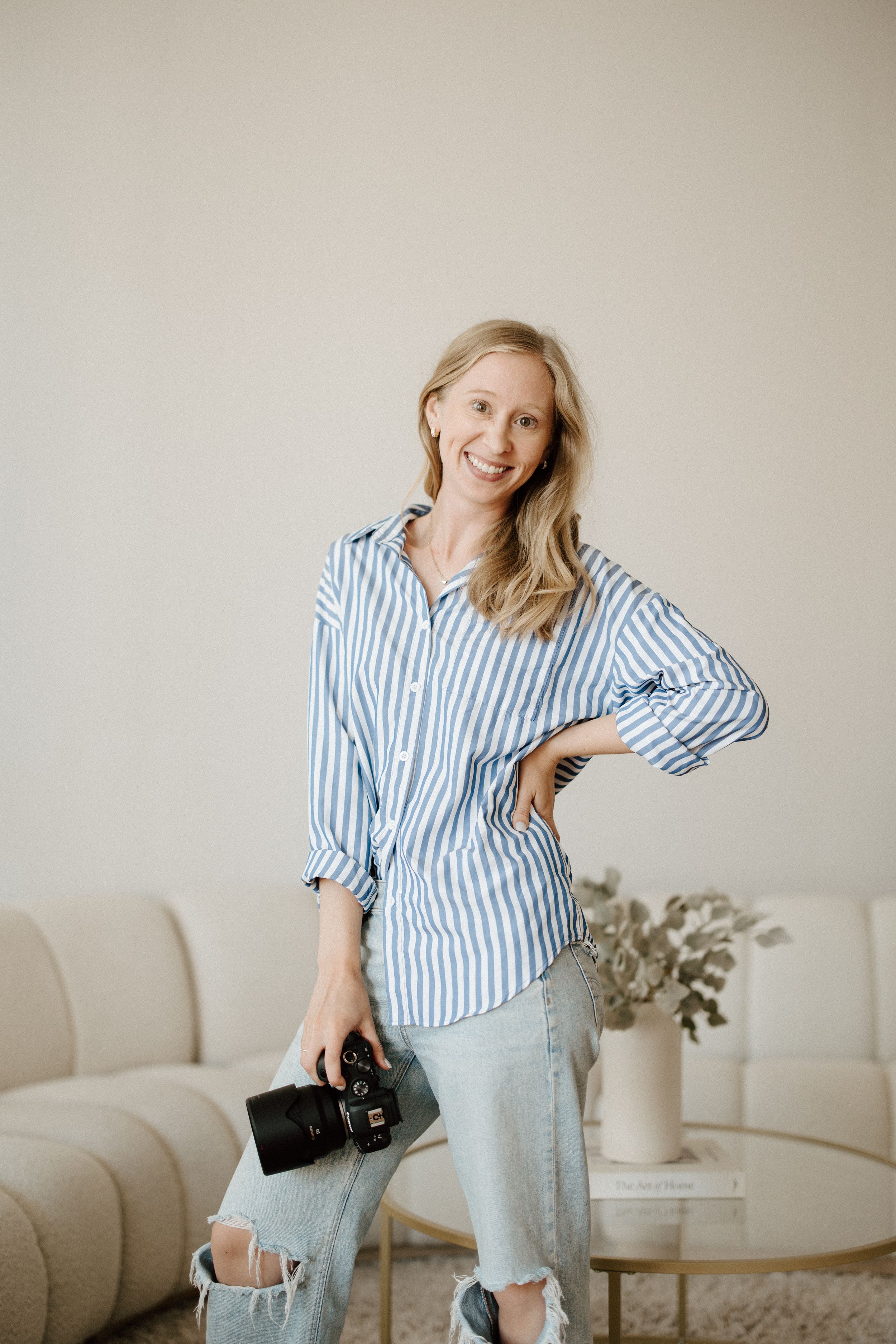 Smiling blonde female photographer holding a camera in a bright modern studio wearing a striped shirt.