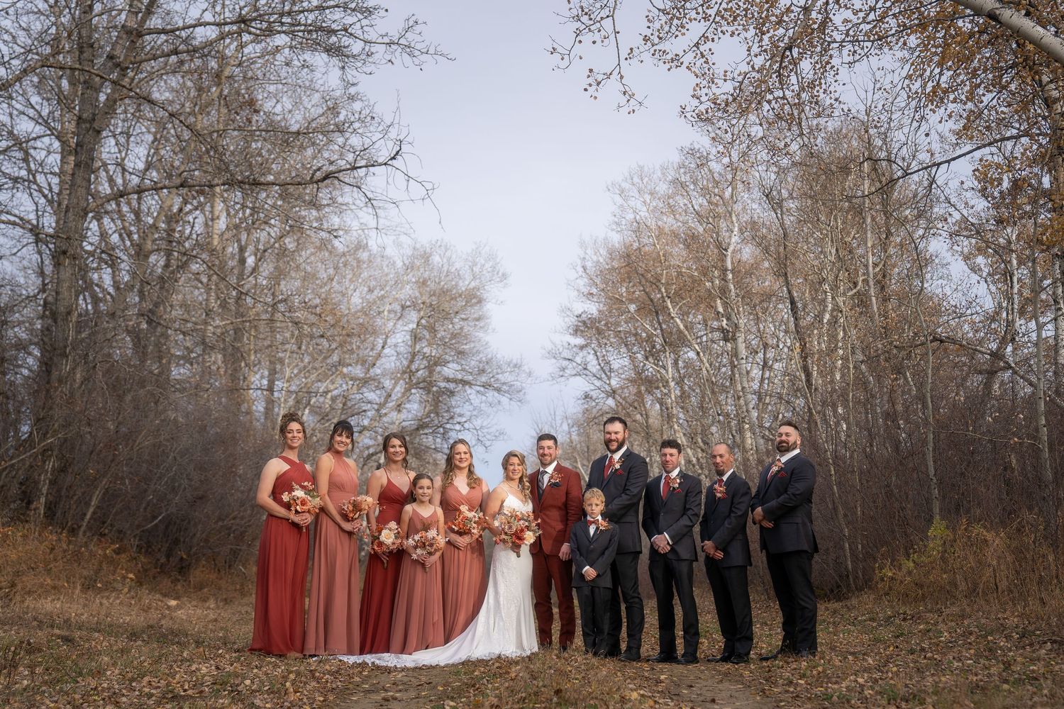 Wedding party stands together in autumn woods with bare trees and fallen leaves creating a rustic outdoor setting.