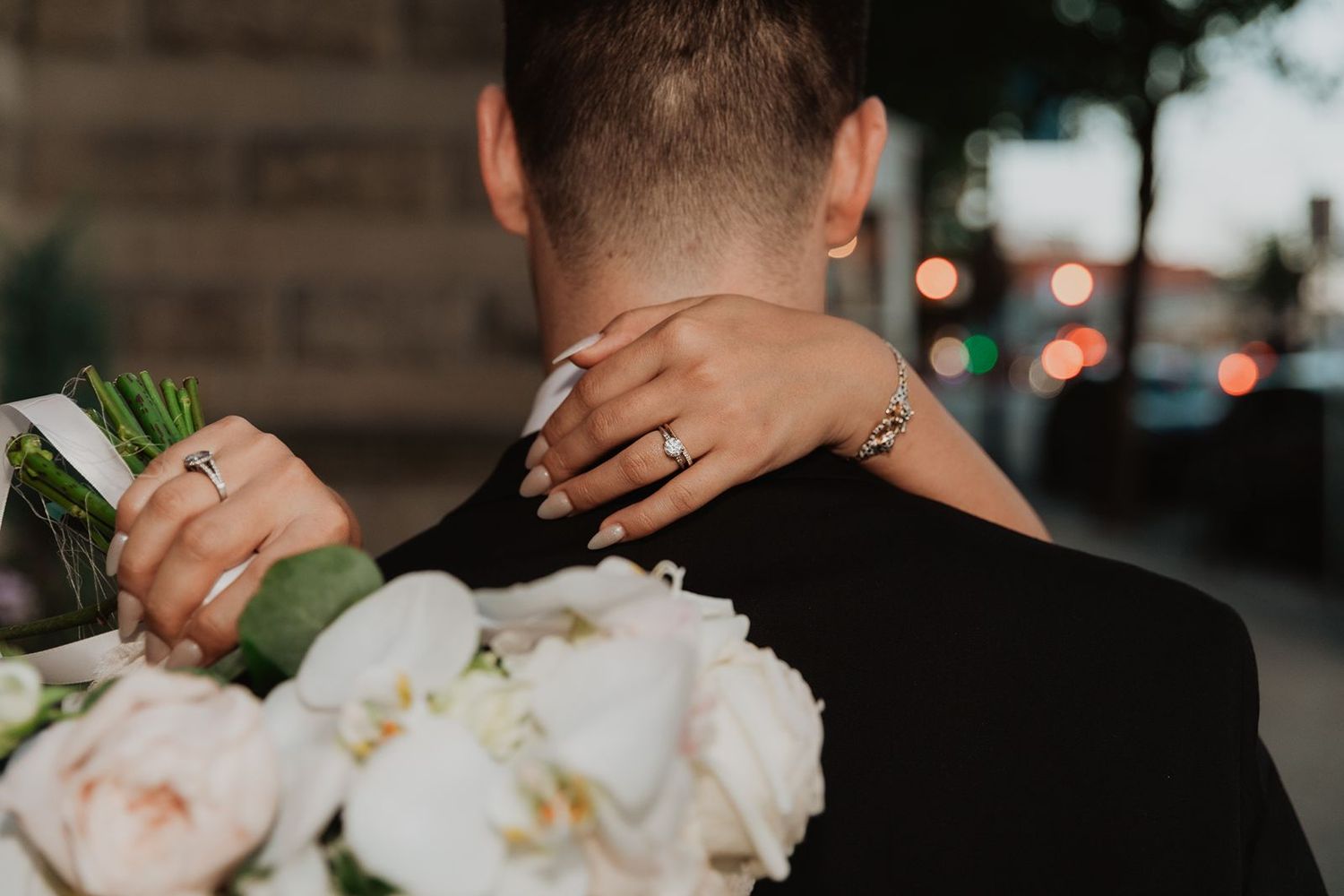 Bride with diamond ring embraces groom from behind holding a white peony bouquet at wedding.