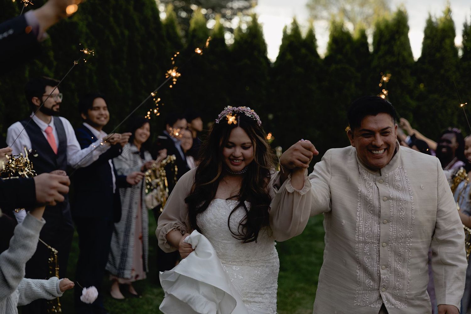 Bride and groom walk joyfully through a sparkler send-off surrounded by cheering wedding guests at night.
