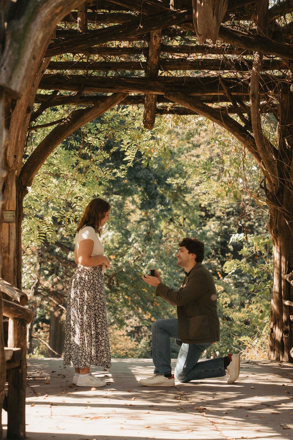 A romantic proposal moment under a rustic wooden pergola surrounded by lush greenery on a sunny day.