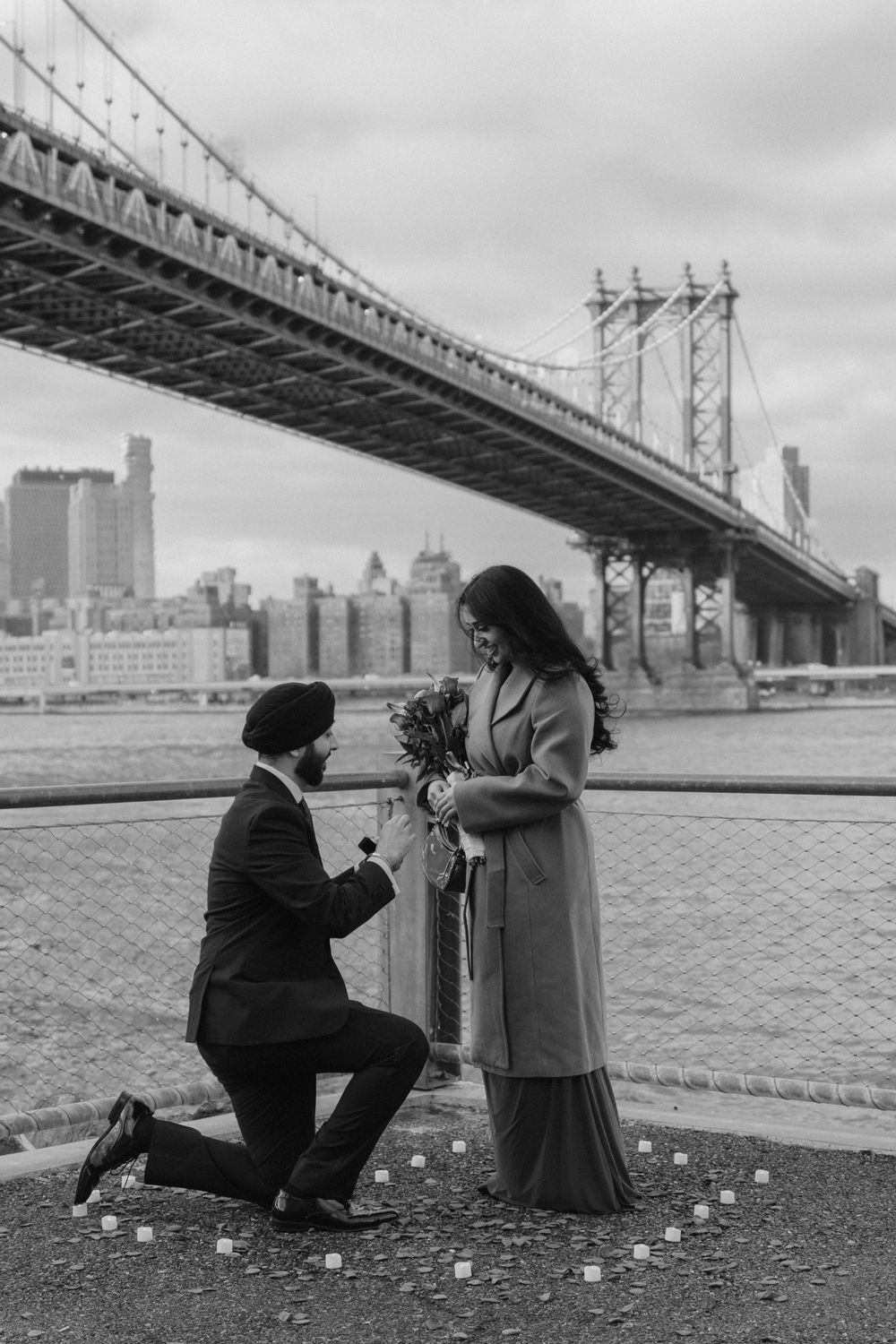 Romantic proposal moment captured under bridge with city skyline and candles creating ambiance.