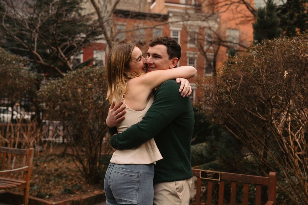 A couple embracing in front of autumn foliage and brick buildings.