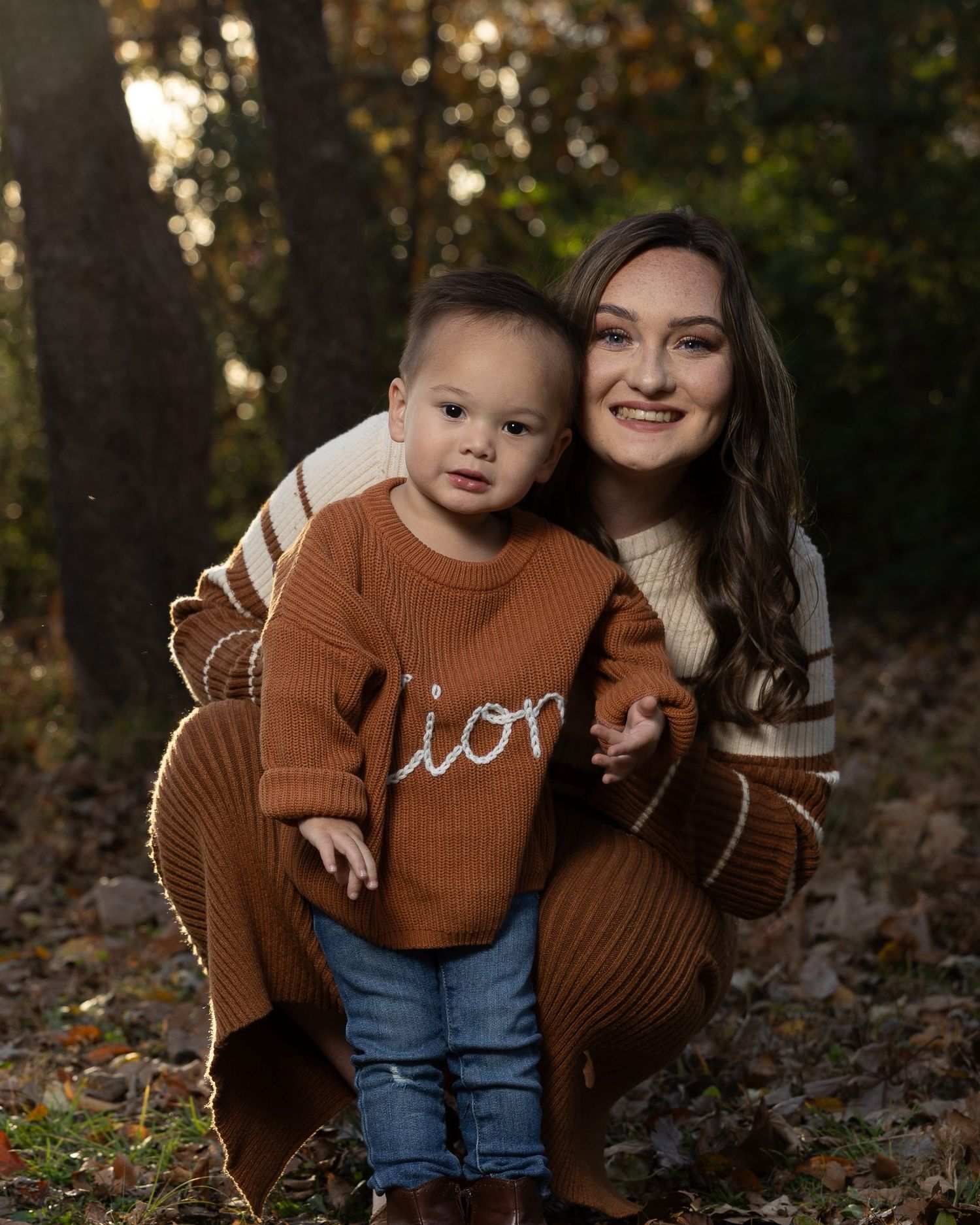 Mother and baby wearing matching brown sweaters pose together in autumn leaves during outdoor family photoshoot.