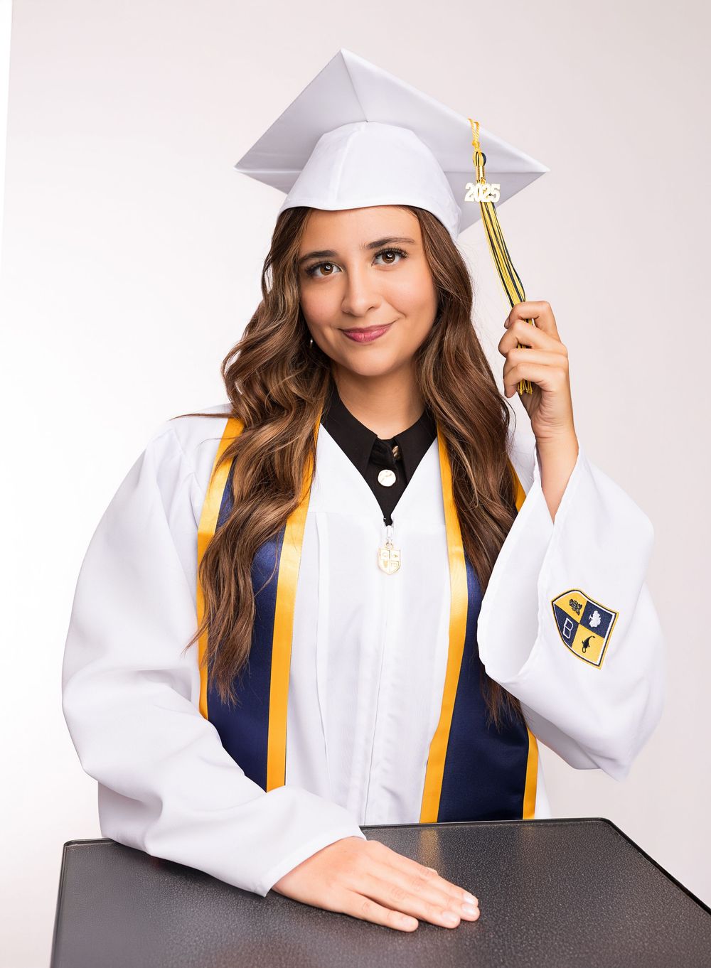 Graduate in white cap and gown with gold tassel poses against light background during graduation photoshoot.