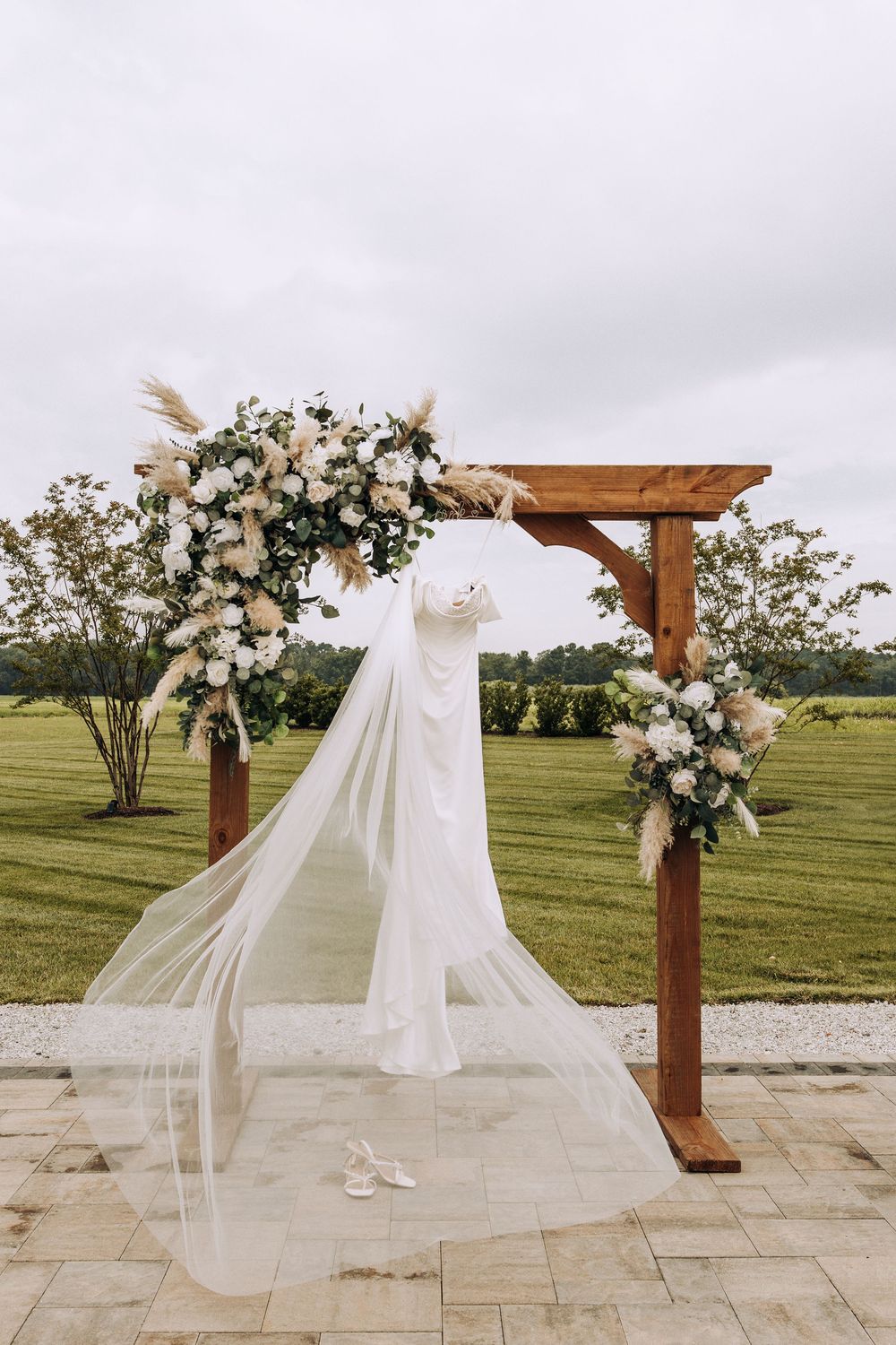 Wedding dress displayed on a wooden floral arch with white roses and pampas grass.