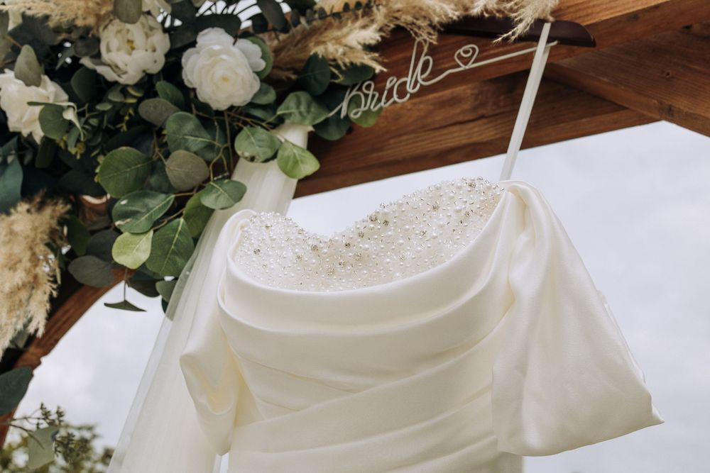 Close-up of a wedding dress hanging on a floral arch with a bride sign.
