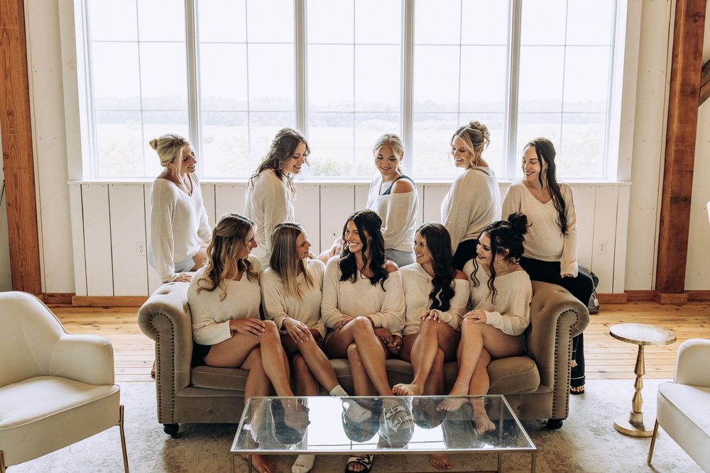 Bridesmaids in matching cream robes gathered together near a large bright window.