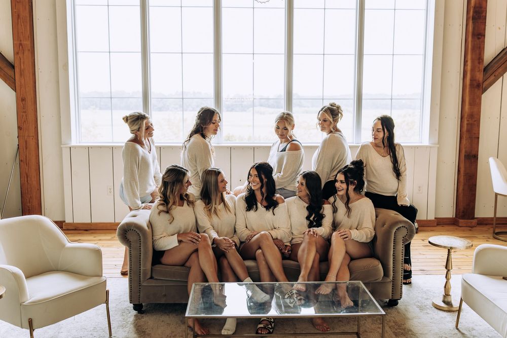 Group of bridesmaids in matching cream robes posing together on a couch near a large window.