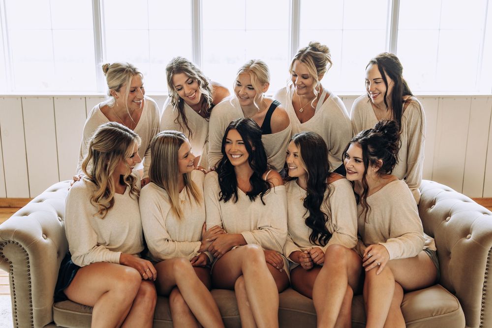 Bridesmaids in cream robes laughing and chatting together on a couch by a window.
